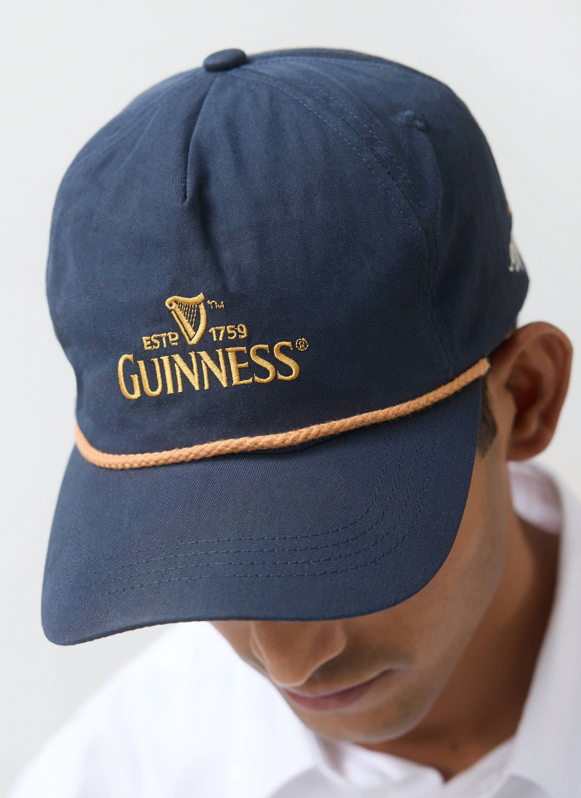 A model wears the navy Guinness x Percival cap, with a close-up on the classic gold Guinness embroidery.