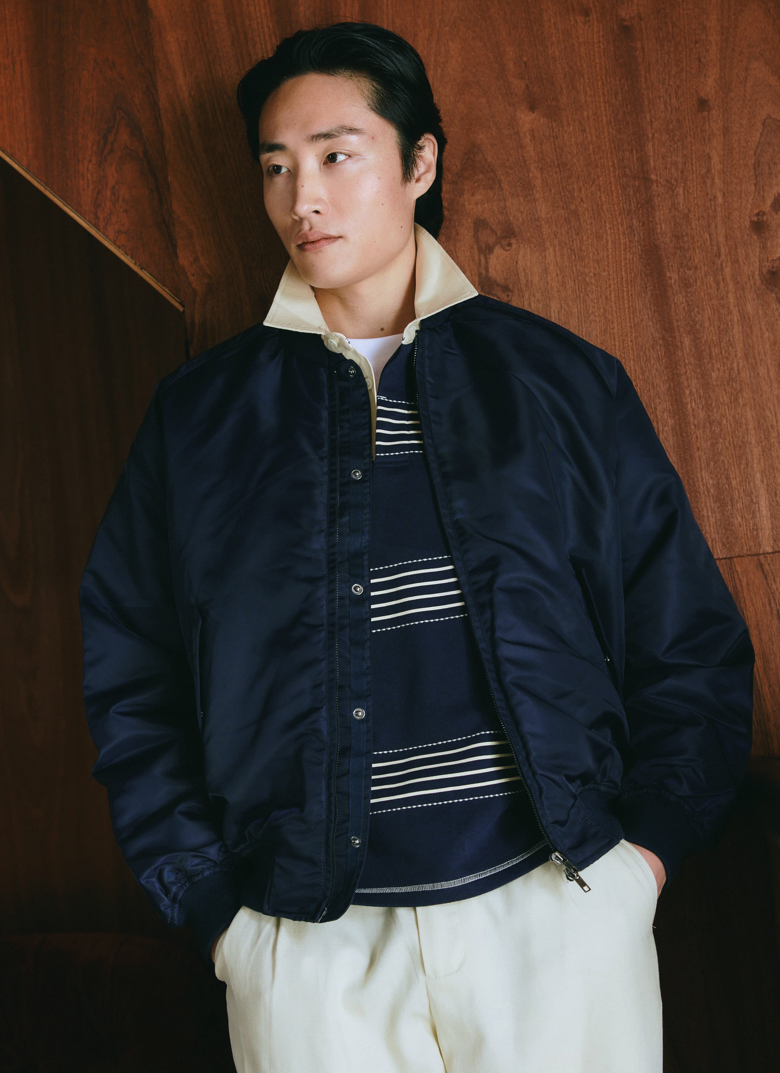 A model wearing the Percival espresso nylon bomber jacket over a striped top against a wooden wall.