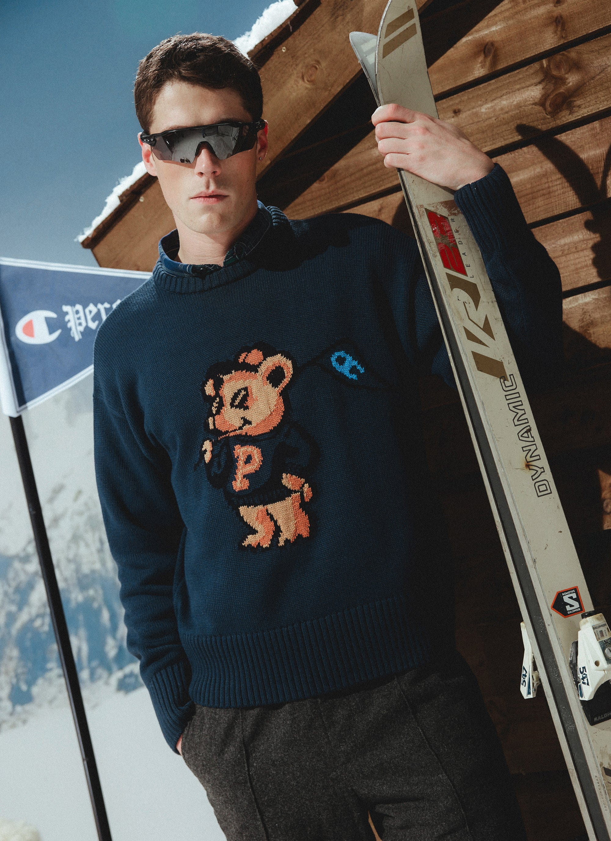 A model wears the navy Champion x Percival Jacquard Bear knit, holding skis in a classic après-ski setting.