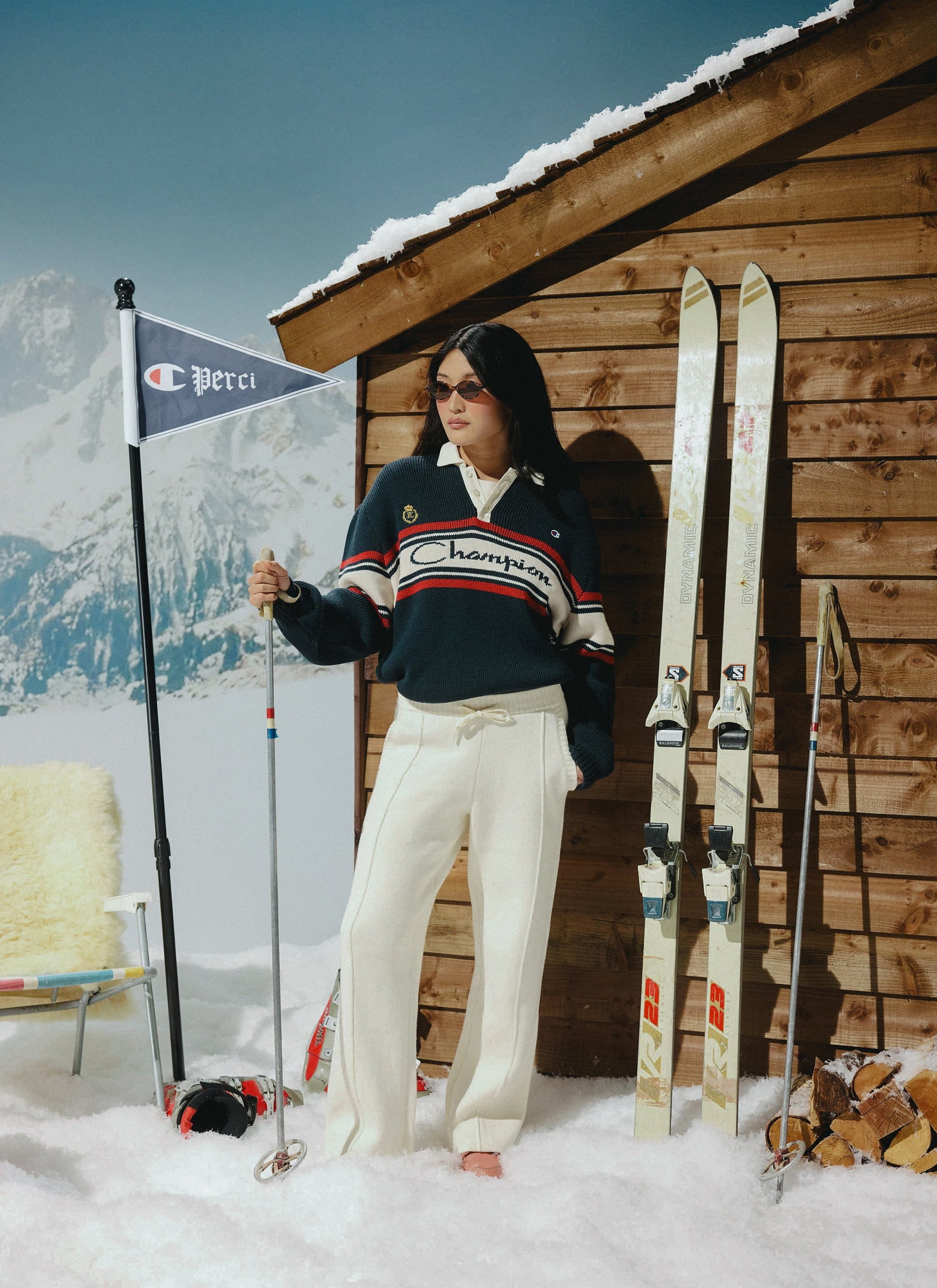 A model wears the Champion x Percival navy knitted rugby shirt in a retro après-ski scene beside a wooden cabin and skis.