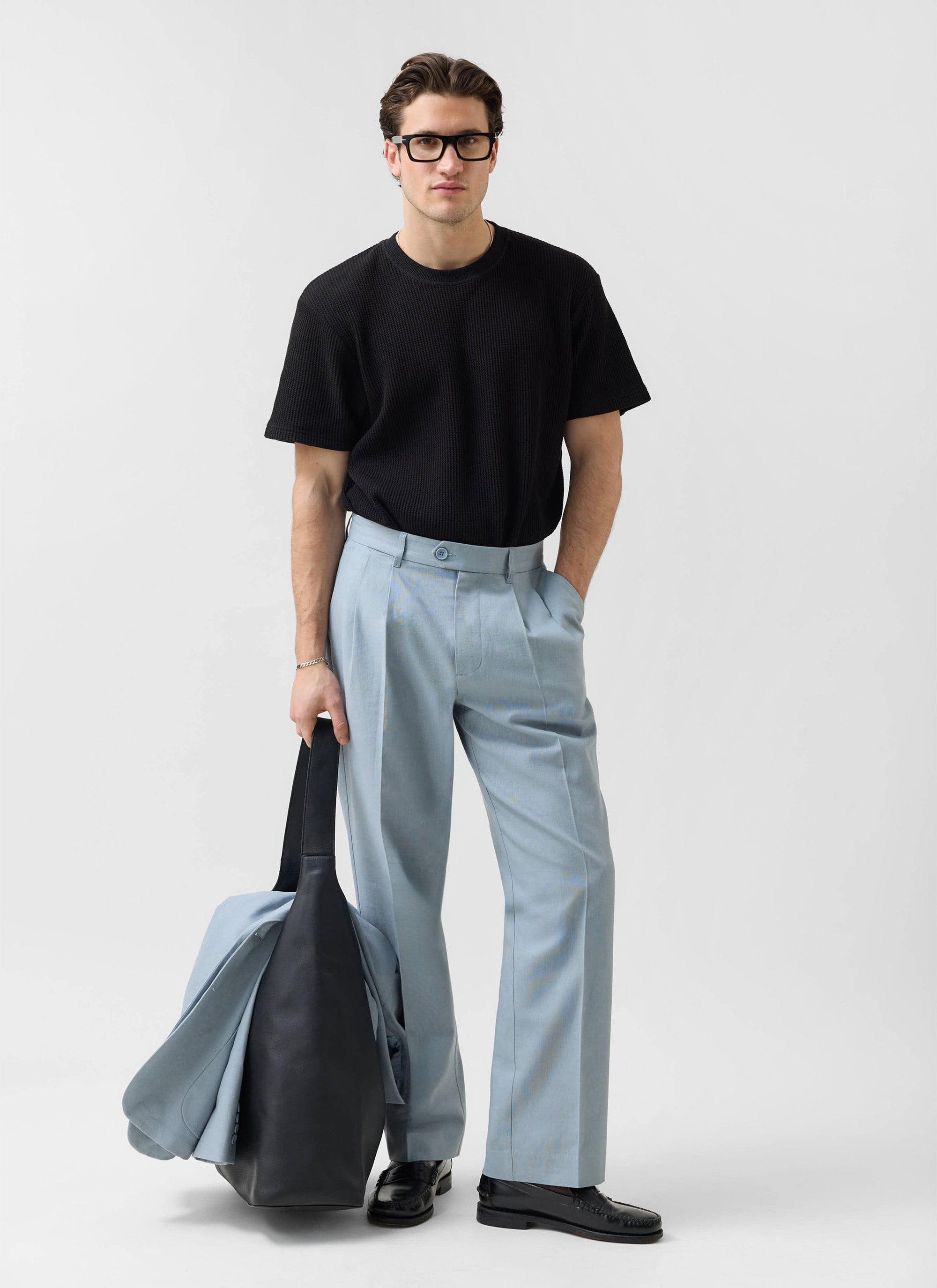 A model wears the Percival light blue linen pleated tailored trousers with a black t-shirt and black loafers.