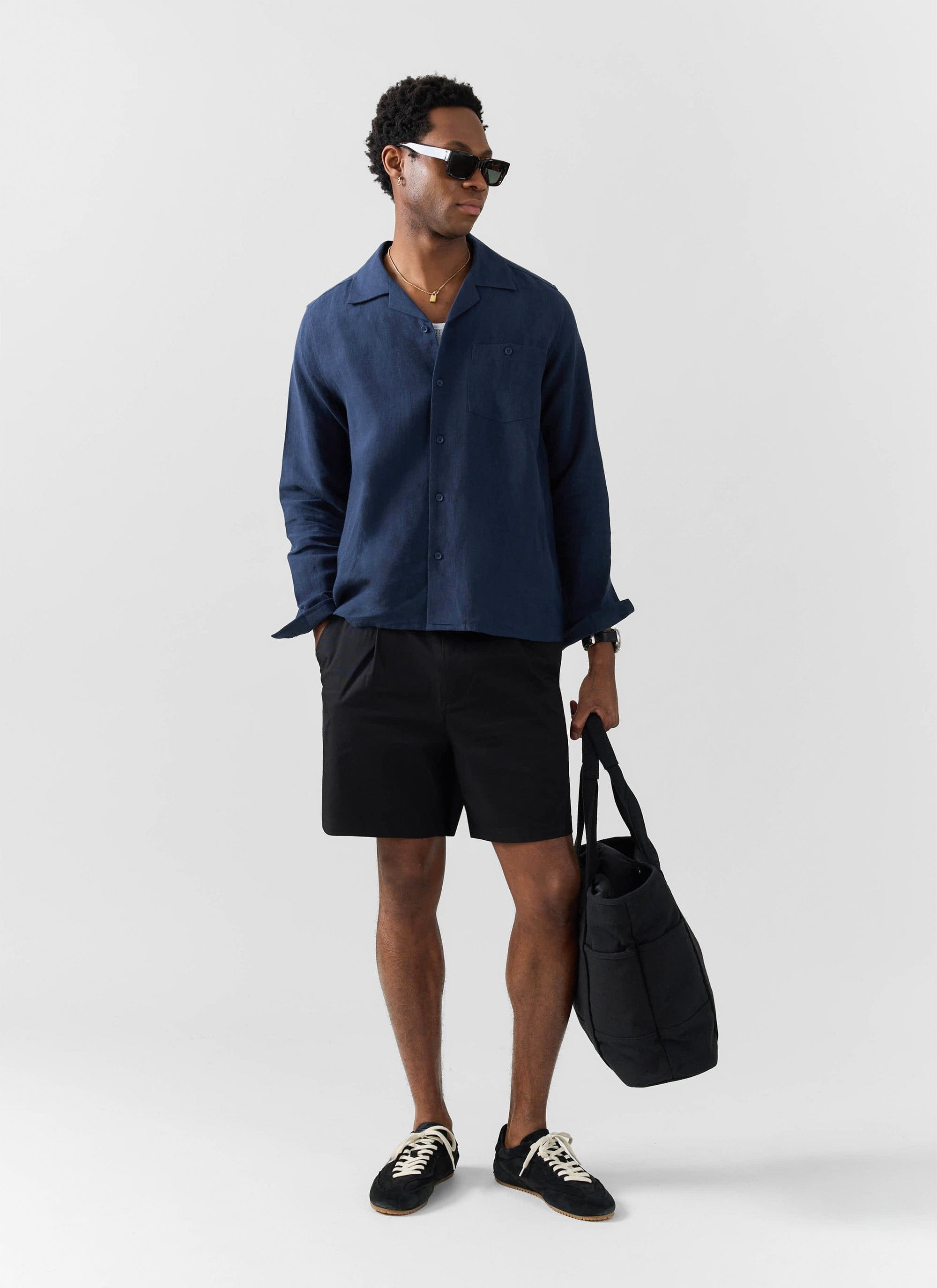 A model wears the Percival Long Sleeve Cuban Linen Shirt in navy, styled with black shorts, sunglasses, and a black tote bag.