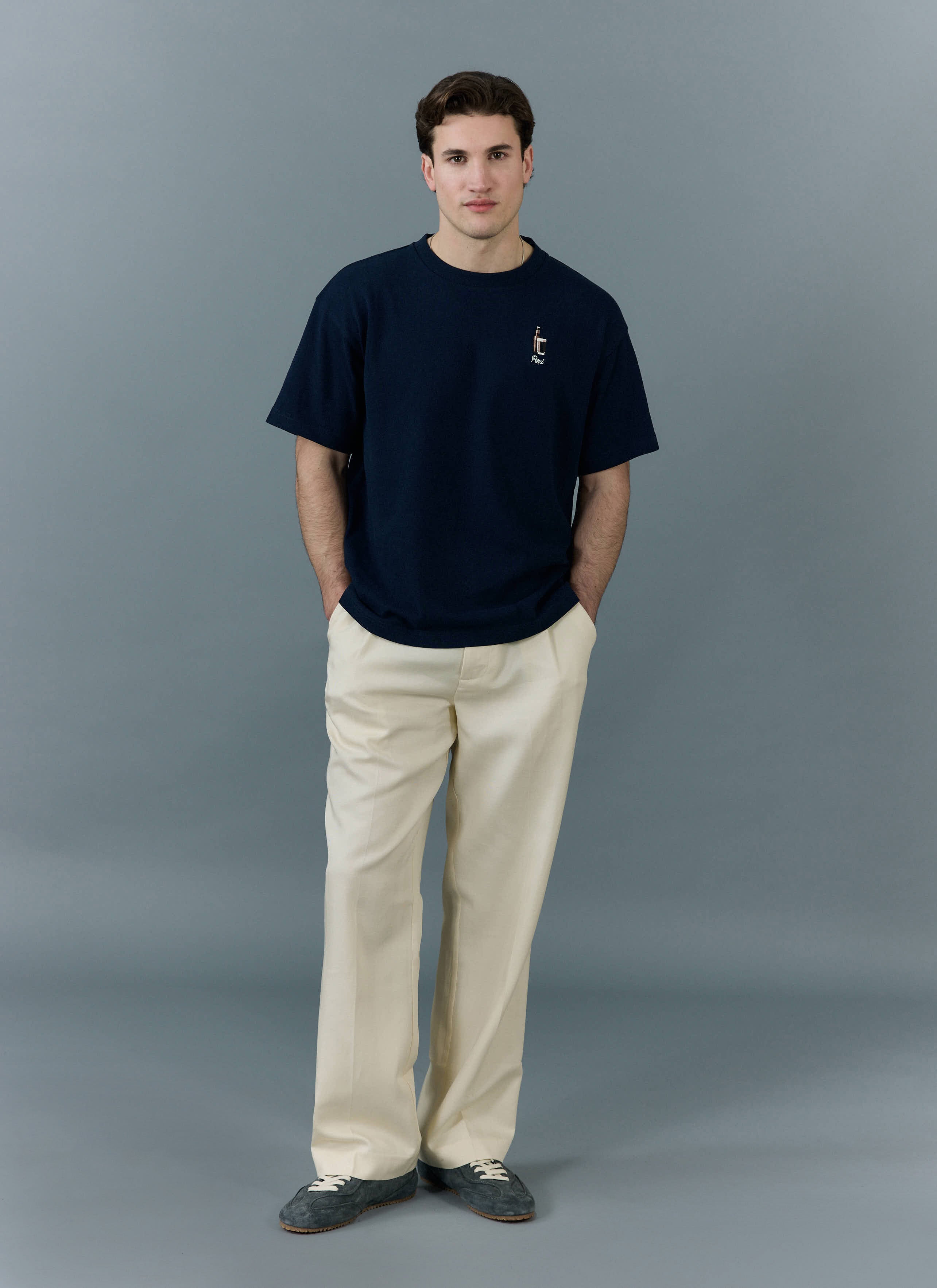 A model wears the Percival Stout Oversized T Shirt in navy, an organic cotton top with stout embroidery, with cream trousers.