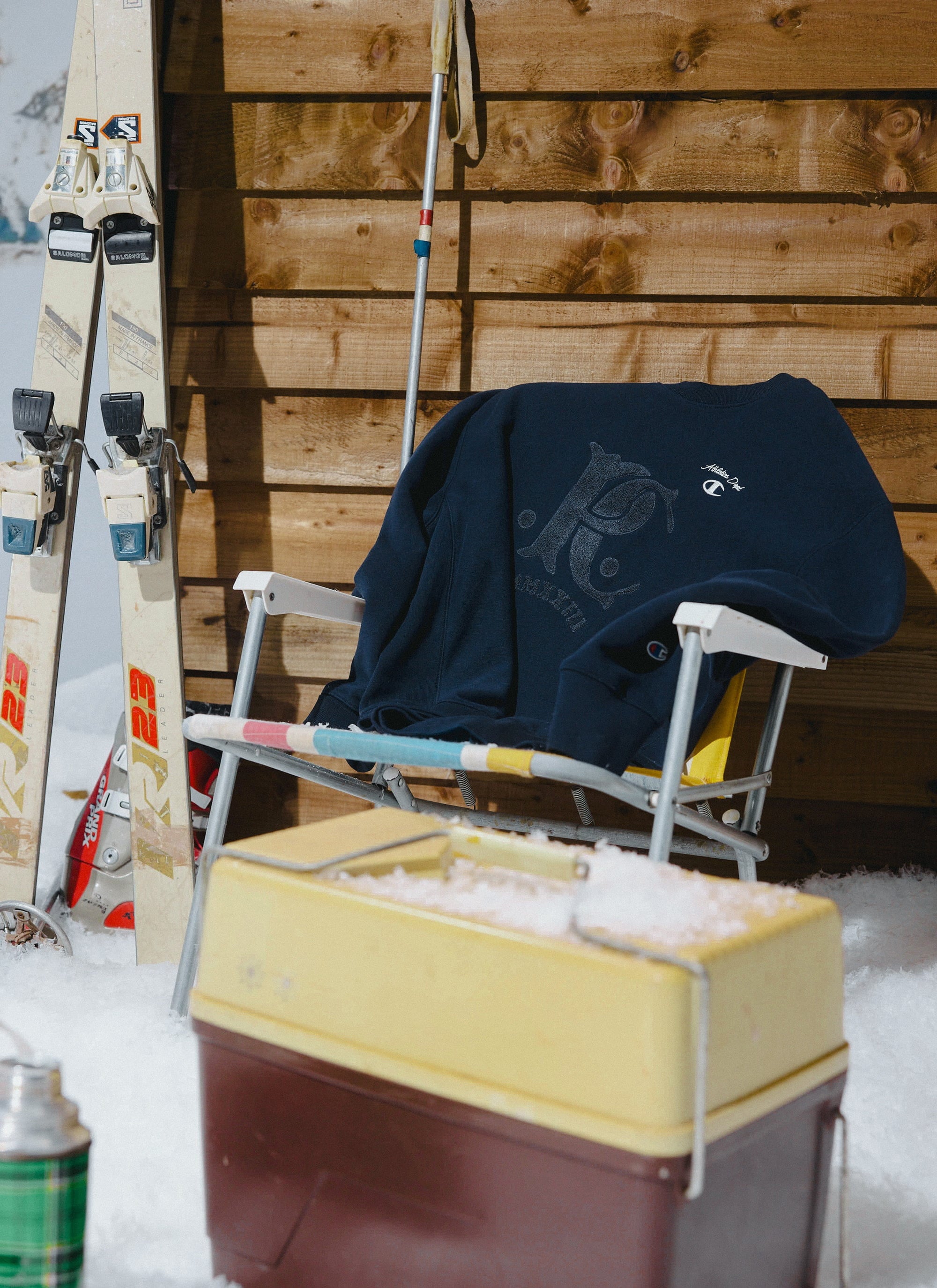 The navy Champion x Percival chenille patch sweatshirt sits on a deckchair in a snowy après-ski scene with vintage skis.