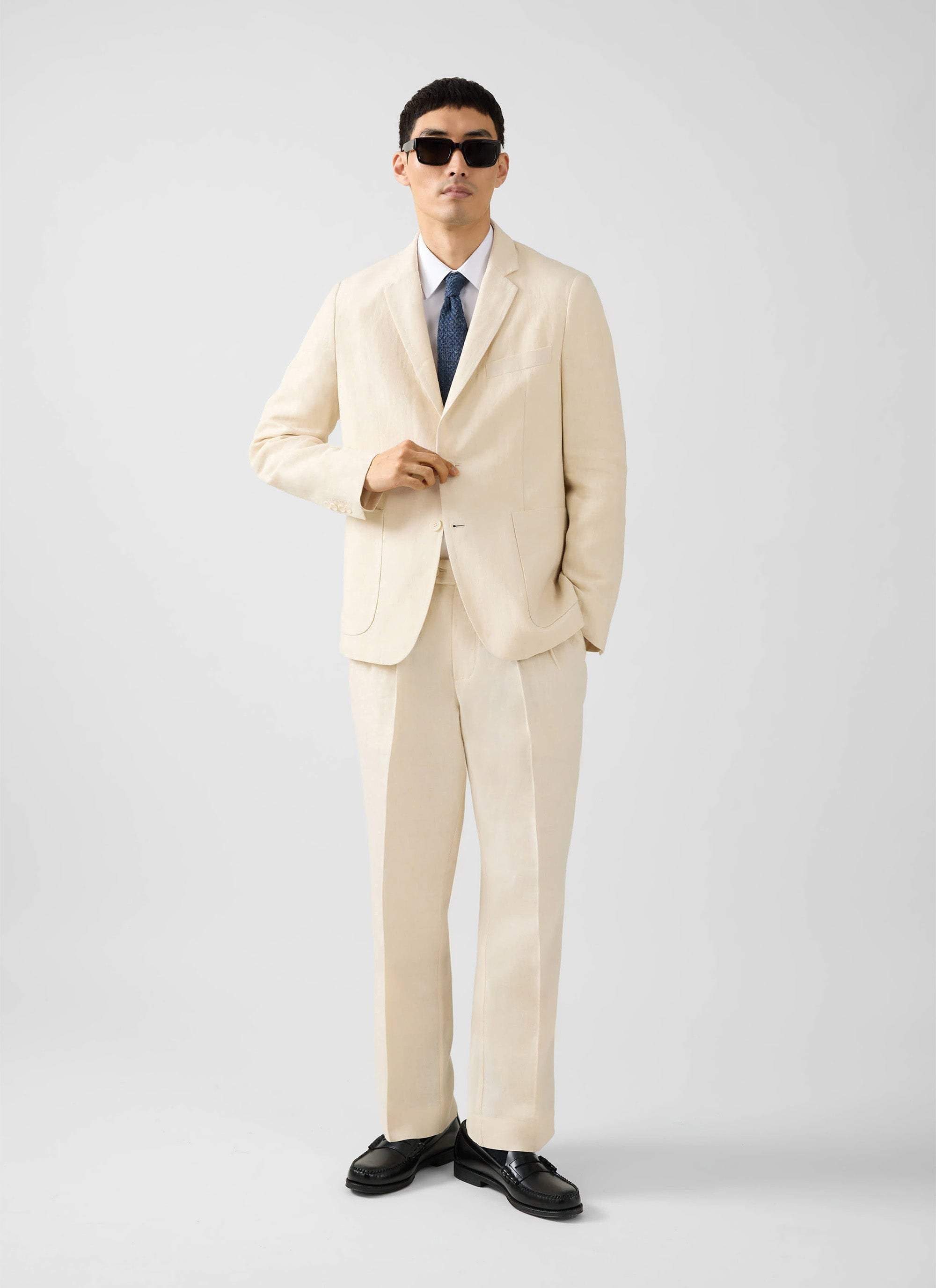 A model wears the Percival cream tailored linen blazer with matching trousers, a shirt, tie, and sunglasses.
