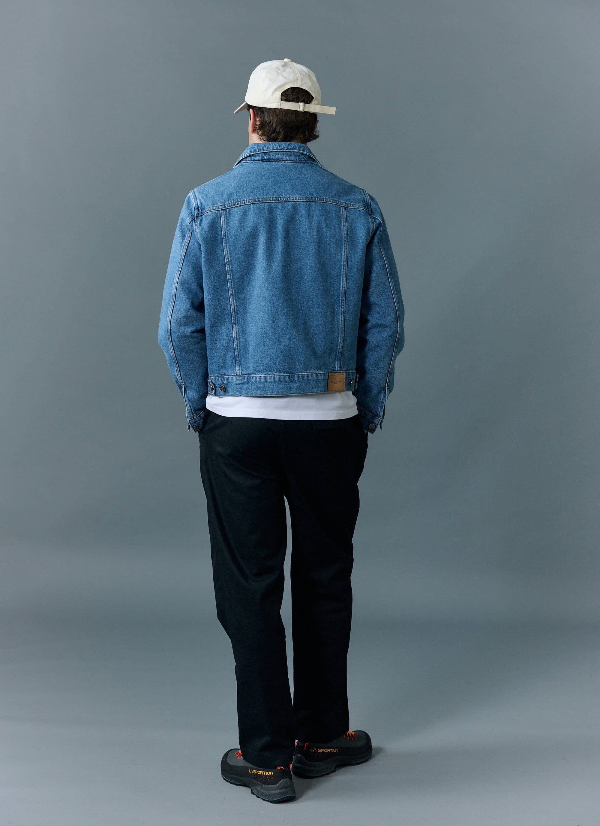 A model shows the back of the Percival Western Trucker Jacket in light wash blue, styled with a white cap and black trousers.
