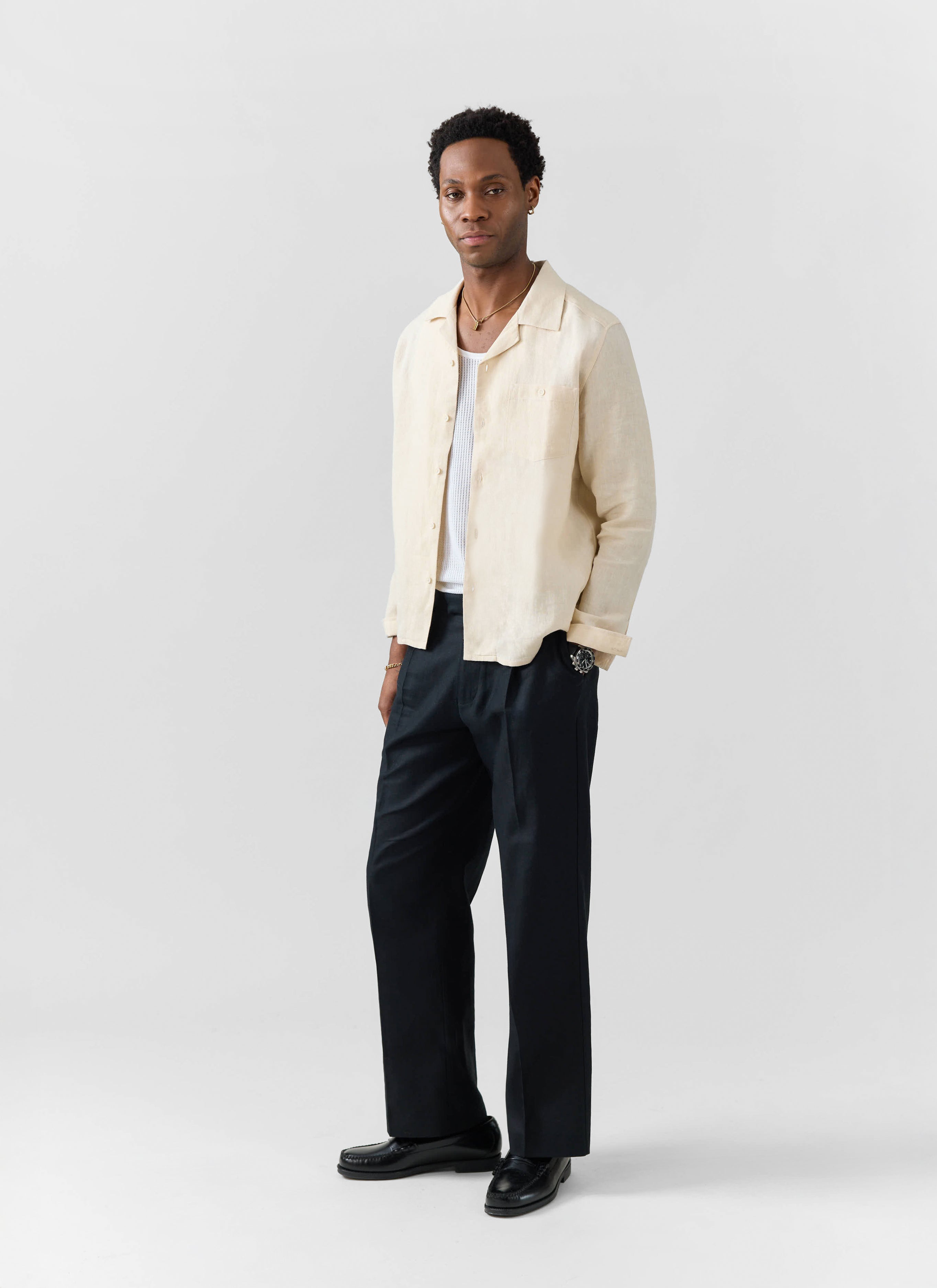 A model wears the Percival cream long sleeve Cuban linen shirt open over a white vest with black trousers and loafers.