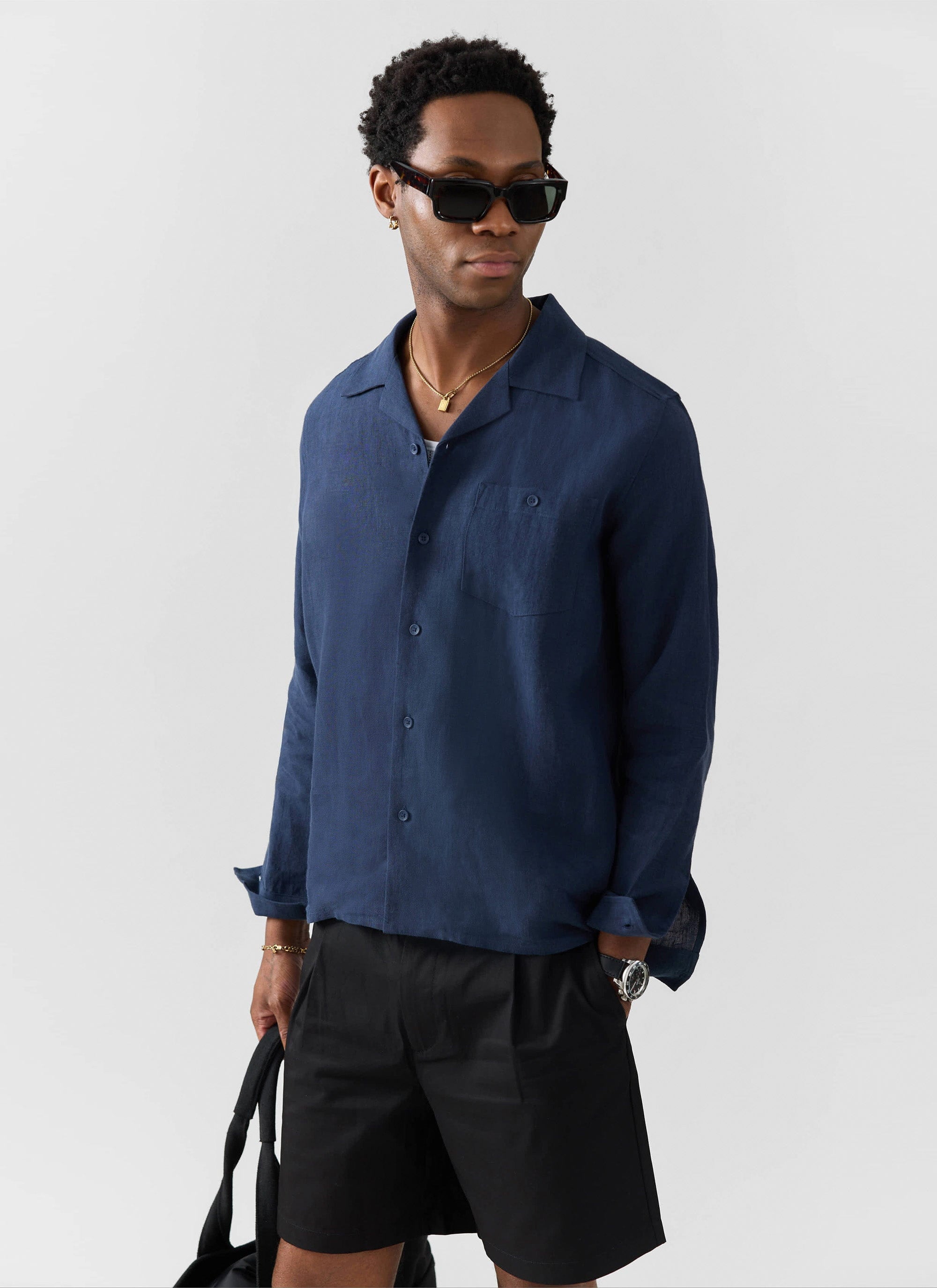 A model wears the Percival Long Sleeve Cuban Linen Shirt in navy with black shorts and sunglasses against a white background.