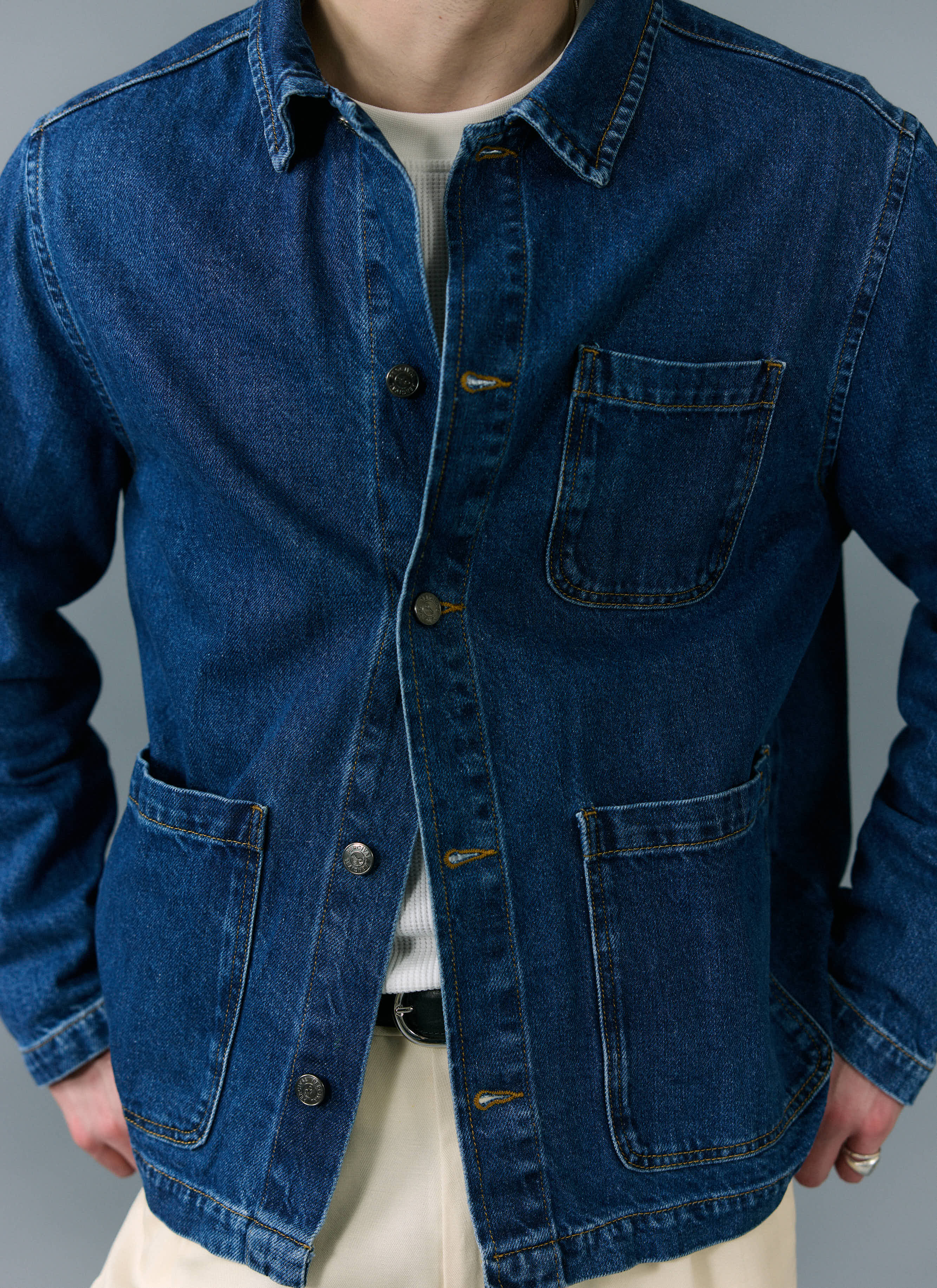 A close-up of a model wearing the Percival Gove Chore Jacket in mid-wash blue denim over a white waffle top.