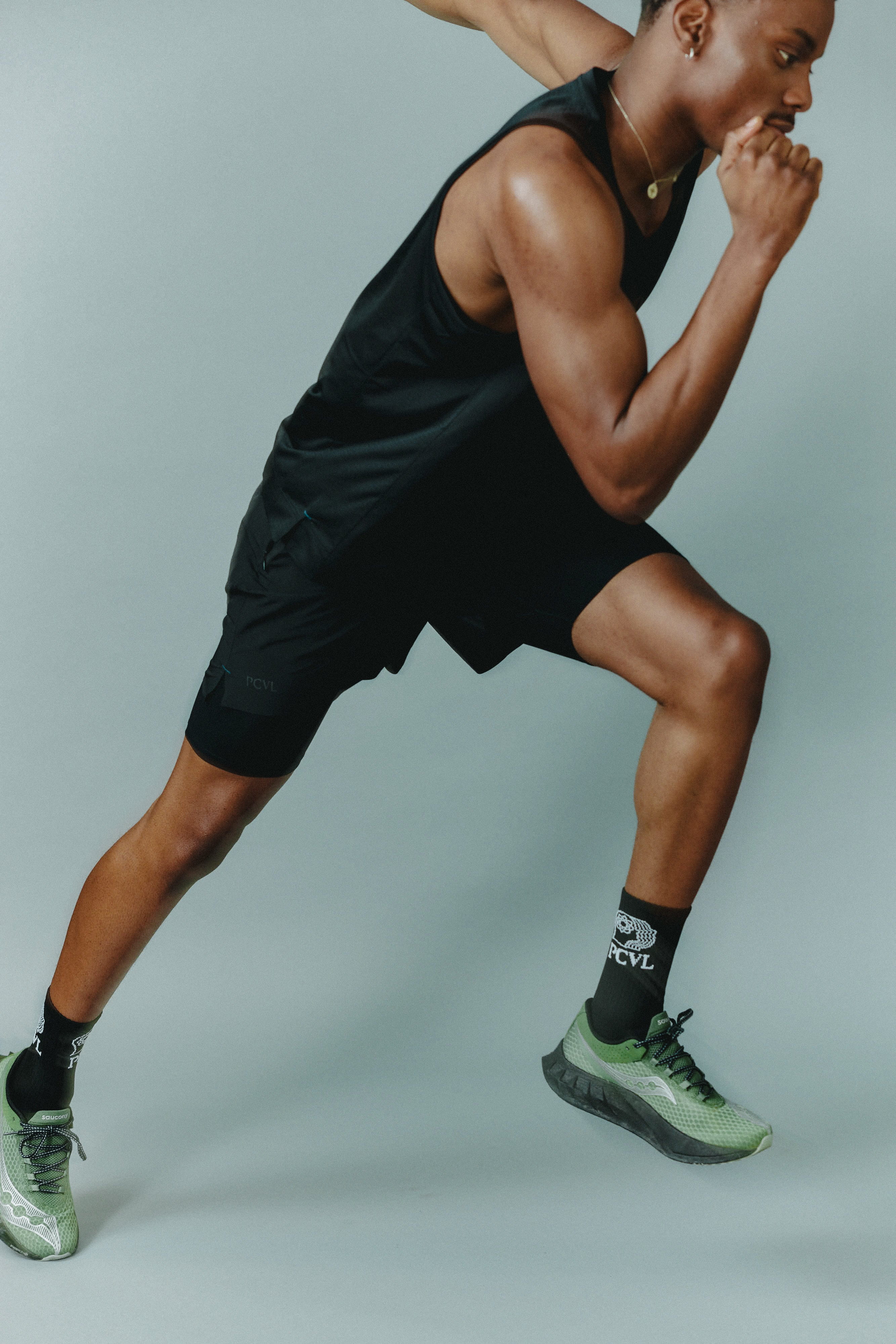 A model in an athletic pose wears the black PCVL Performance Shorts and Sport Socks by Percival against a grey background.