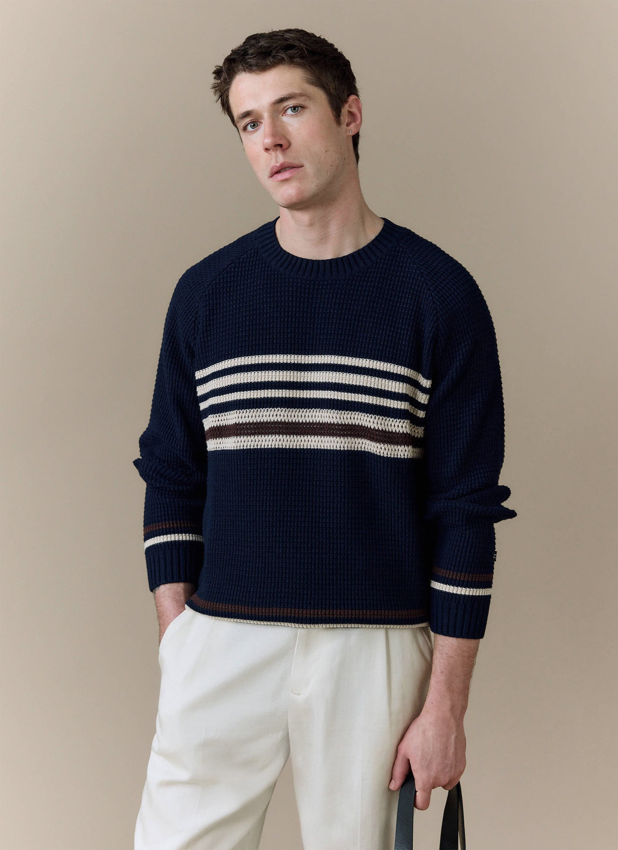 A model wears the navy Sergio Crochet Jumper from Percival, styled with off-white trousers against a neutral background.