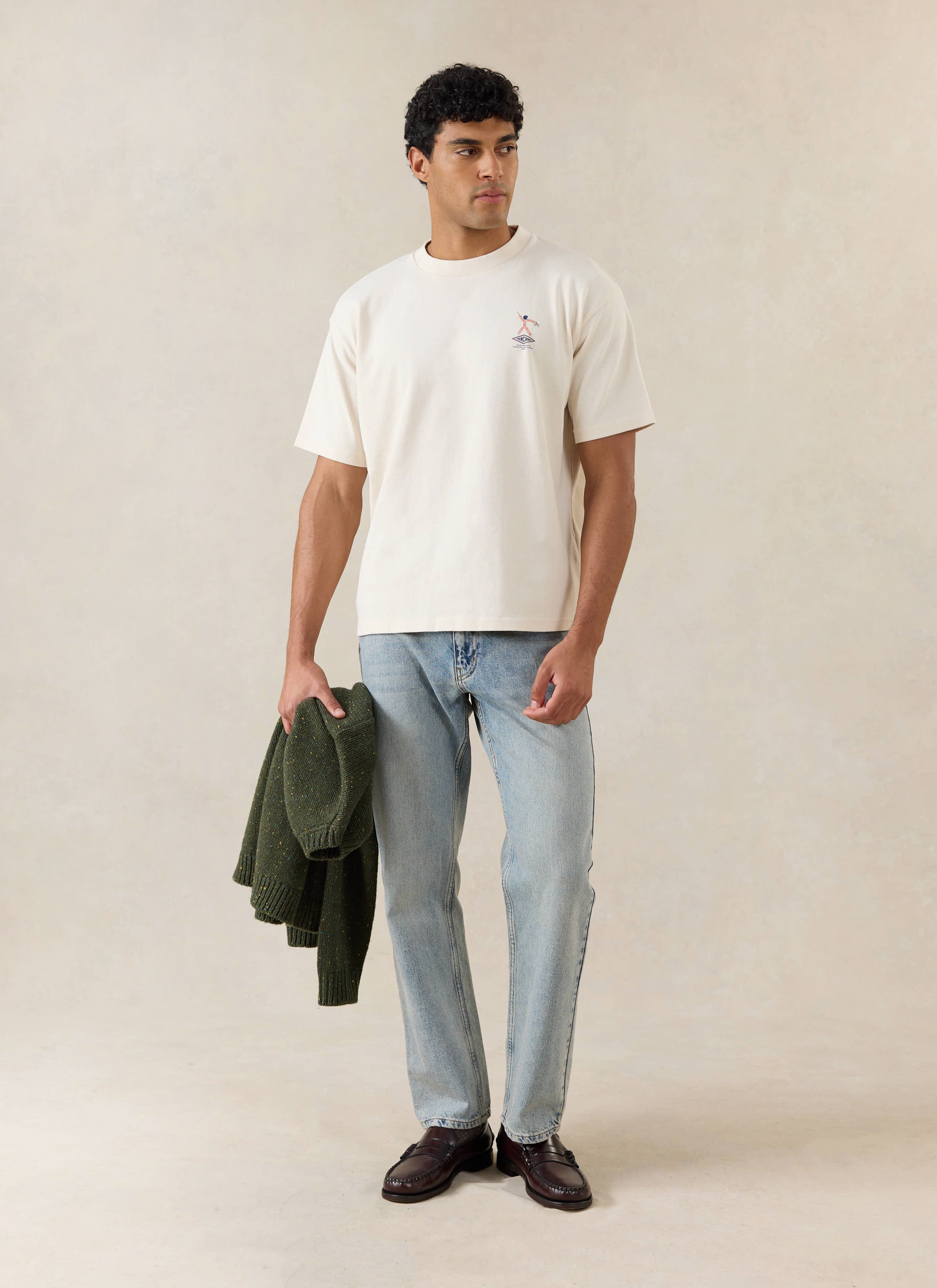A model wears the cream Percival Kingston t-shirt with light jeans and a green jumper.