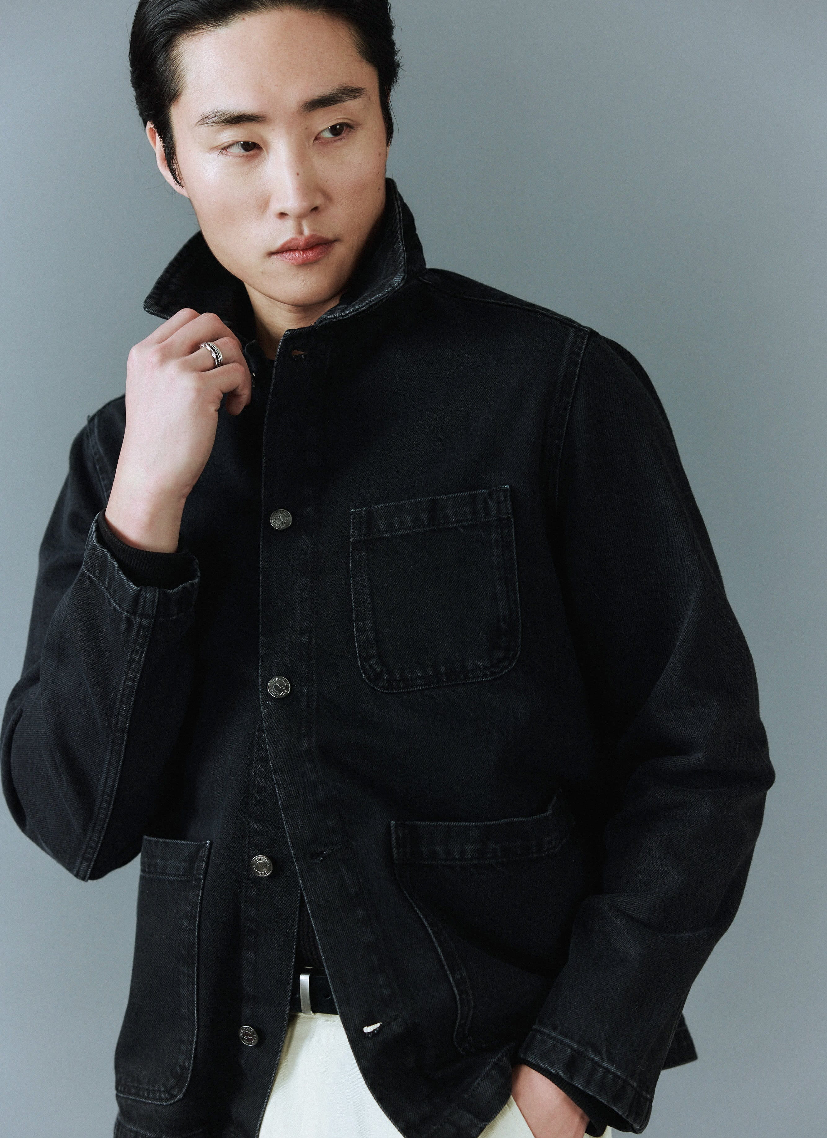 A model styles the Percival Gove Chore Jacket in black denim, holding the collar up against a grey studio background.