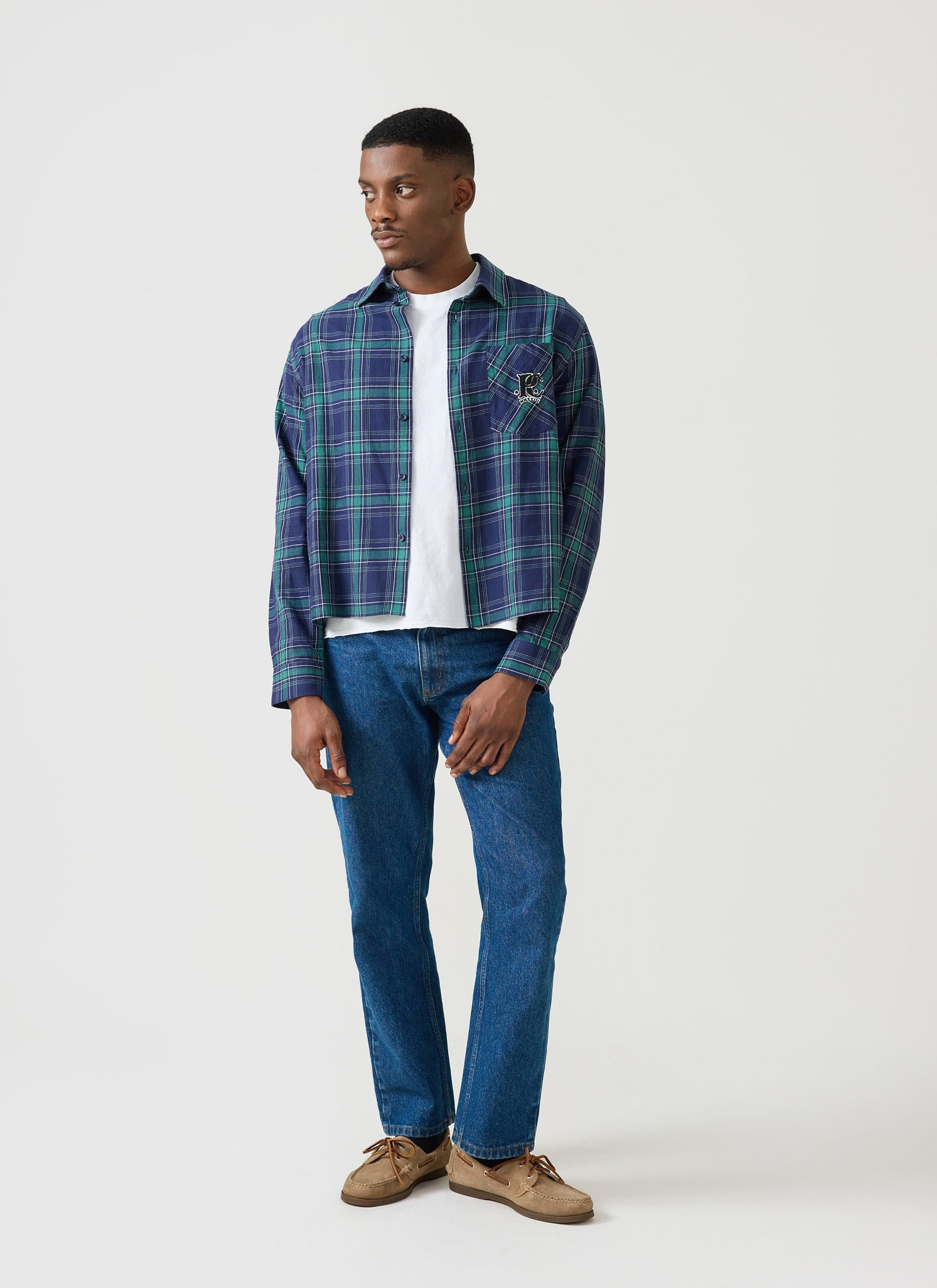 A man models the Percival x Champion check boxy shirt, worn open over a white t-shirt with blue jeans.