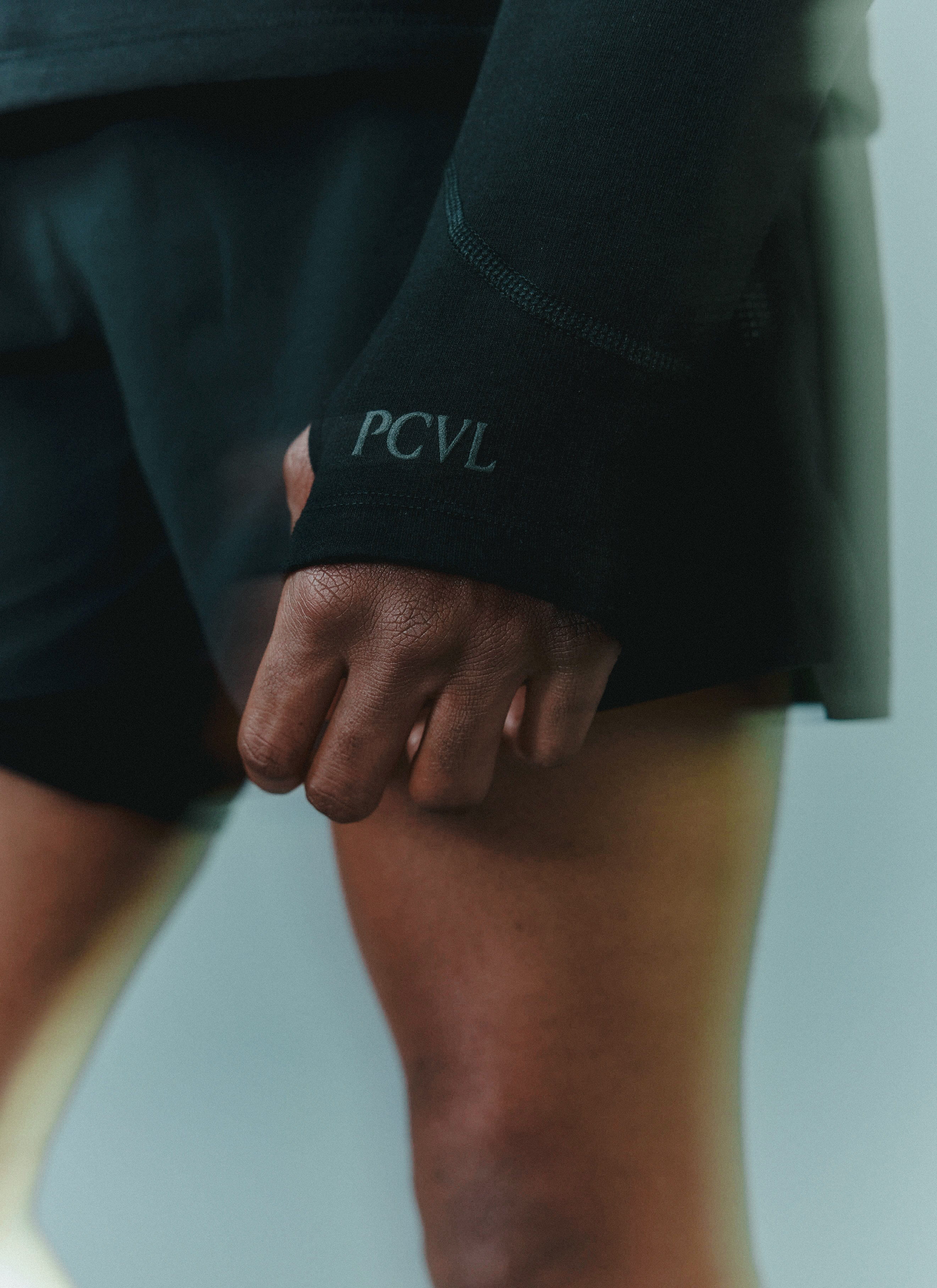 A close-up of a model wearing the PCVL Performance Shorts in black by Percival, with a focus on the tonal PCVL sleeve logo.