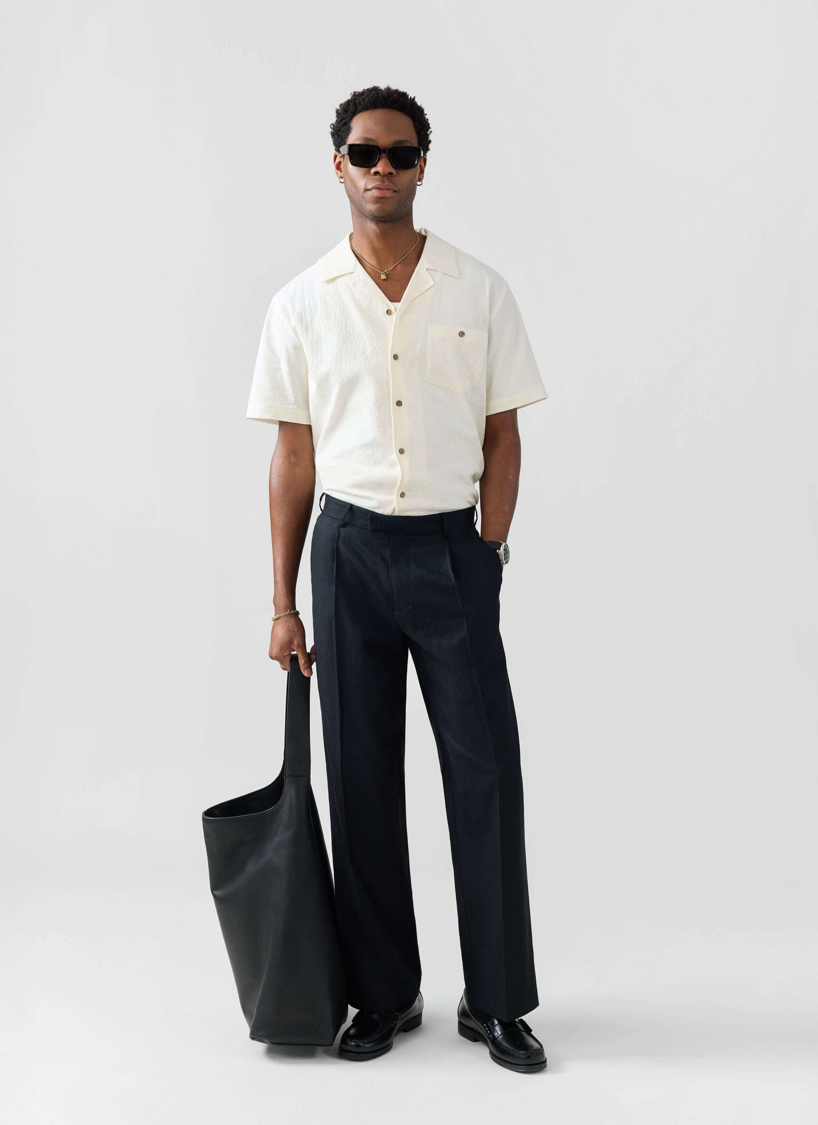 Model wears the Percival ecru seersucker Cuban shirt with wide-leg black trousers, sunglasses and a black tote bag.