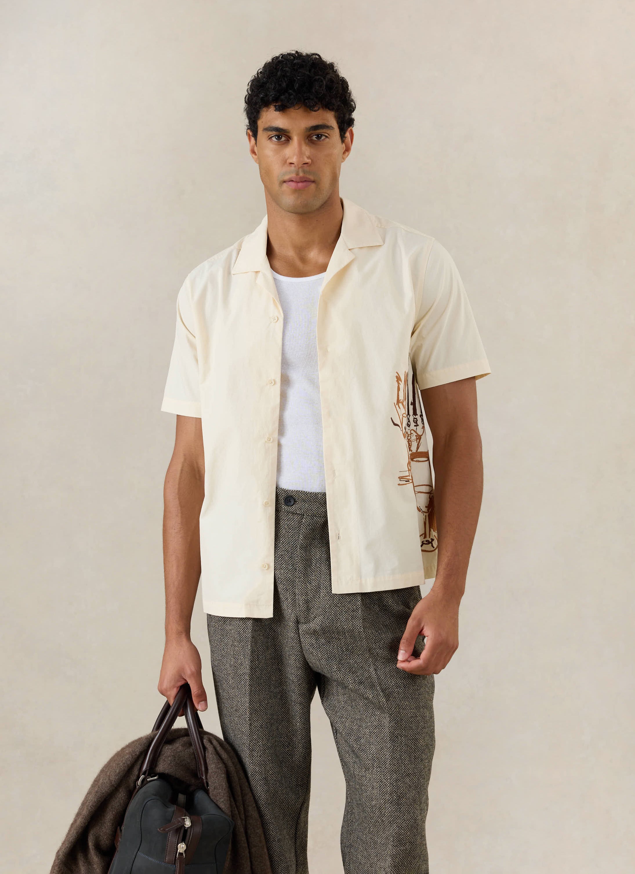 A model wears the Percival Supper Club Cuban shirt in cream cotton with side embroidery, styled with grey trousers.