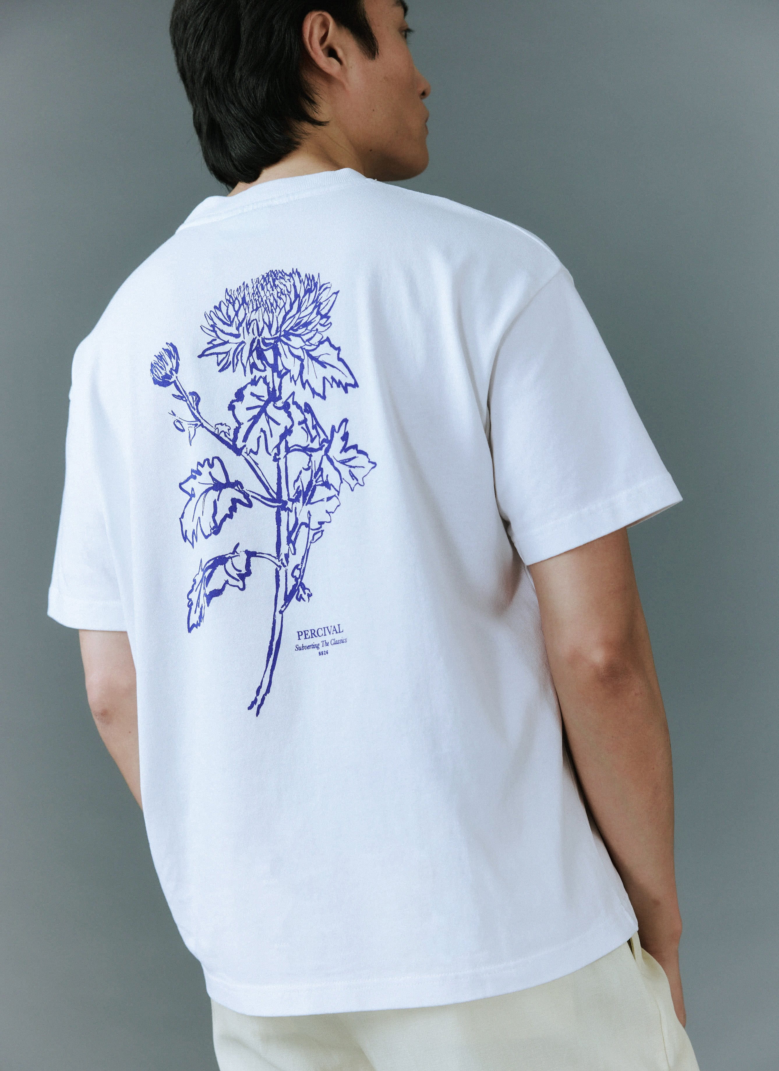 A model seen from behind wears the white Smith Floral t-shirt from Percival, showcasing the blue floral back graphic.