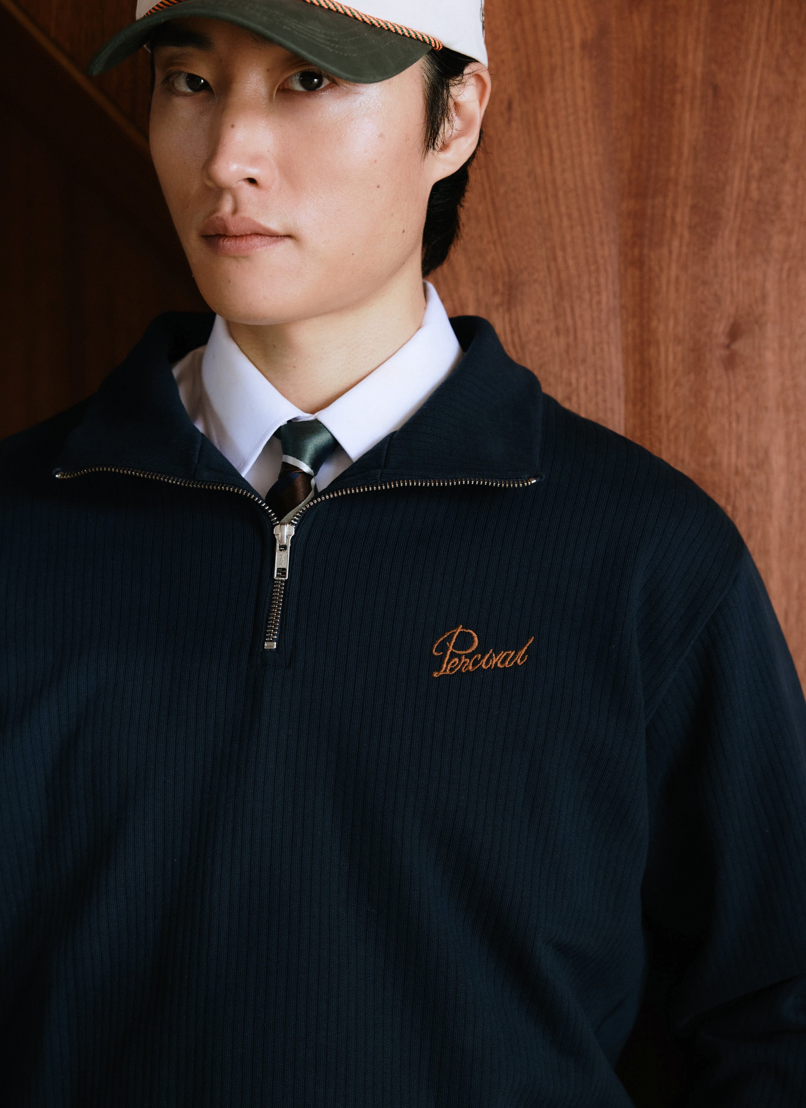 Close-up of a model wearing the navy cotton Percival Conrad funnel neck 1/4 zip jumper with a white shirt and cap.