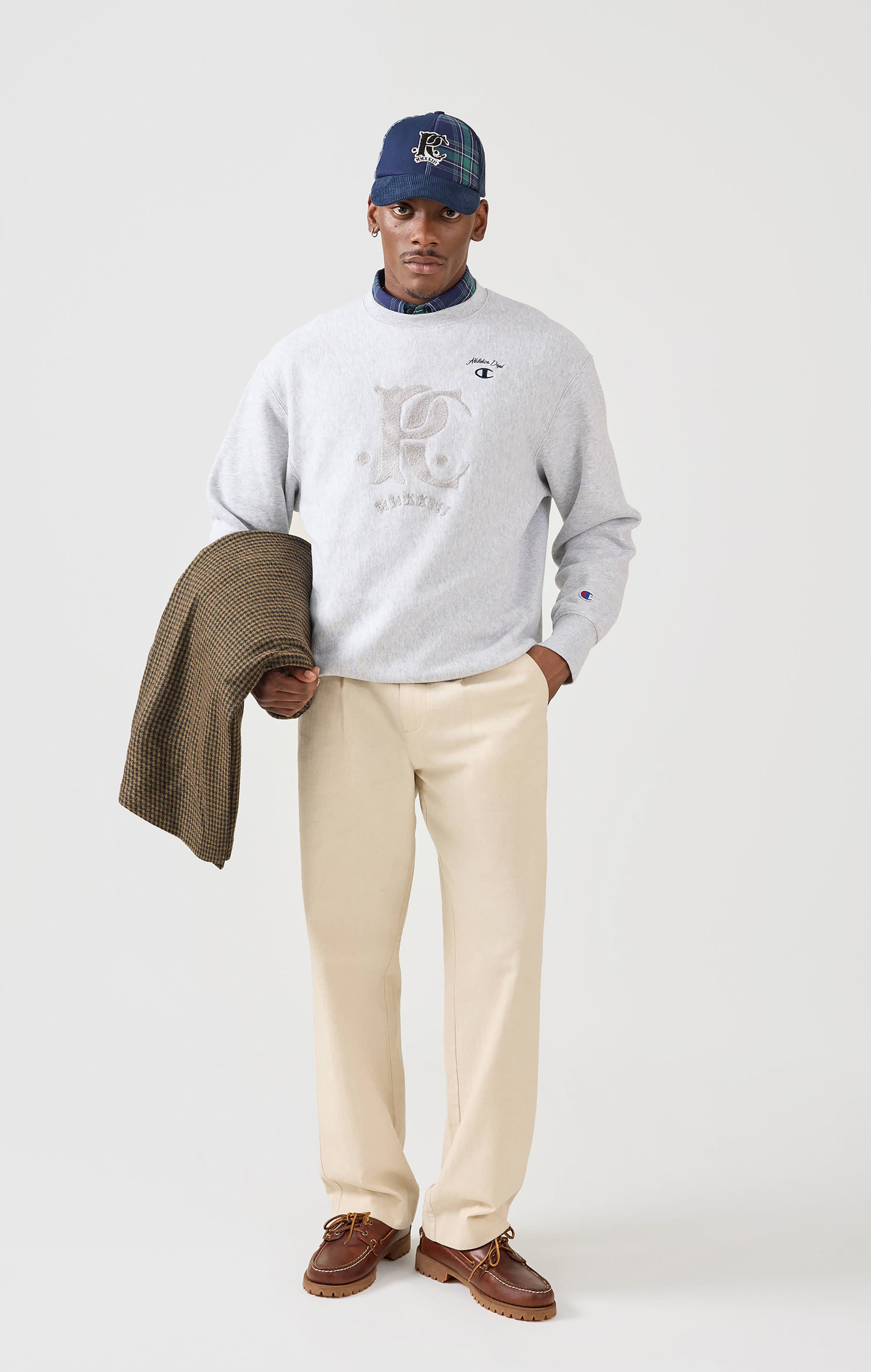 A model wears the Champion x Percival grey Chenille Patch Jersey Sweatshirt, styled with cream trousers and a tartan cap.