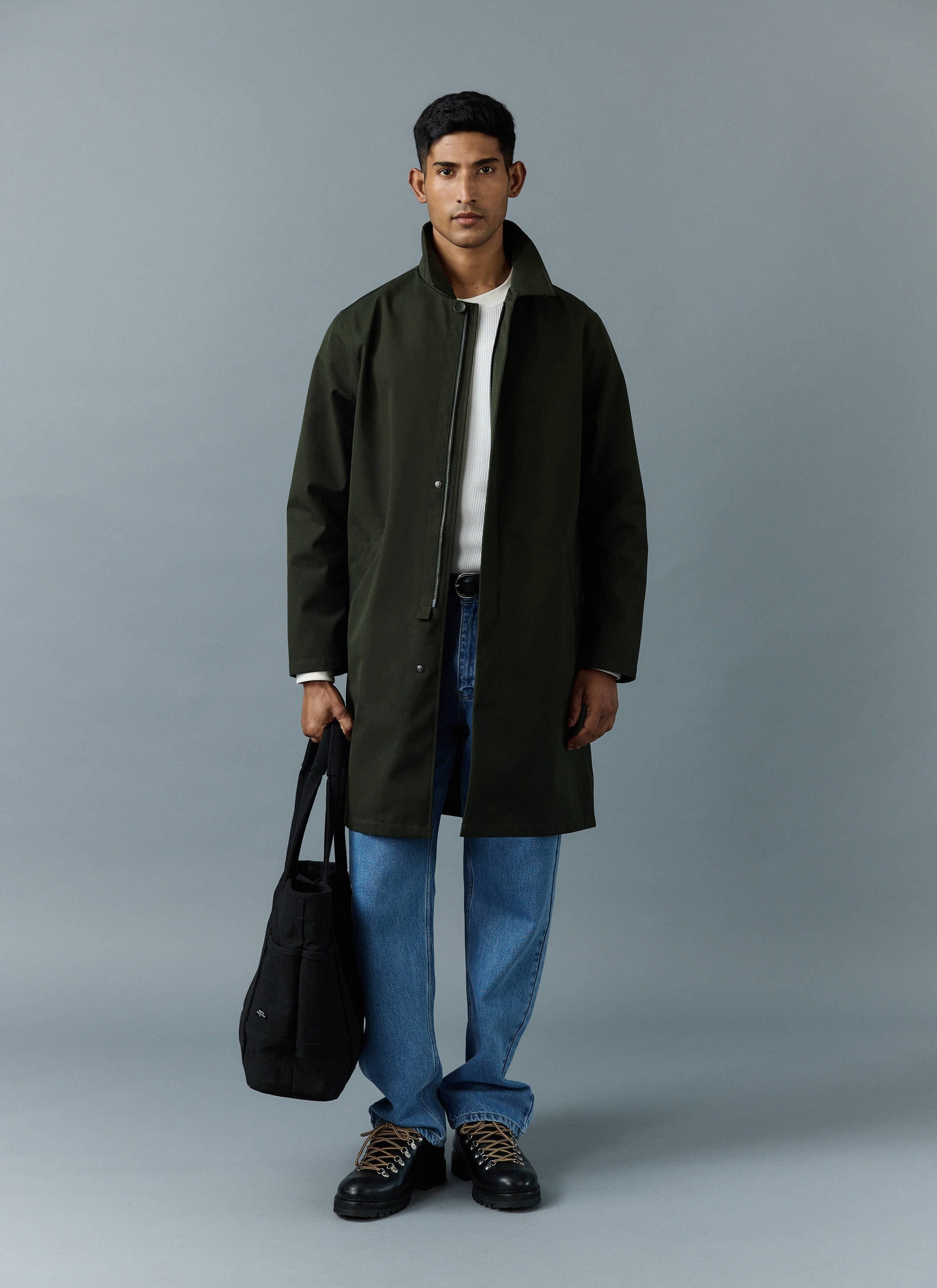 A model wears the Percival Greyson Long Trench Coat in forest green, styled with blue jeans and carrying a black tote bag.
