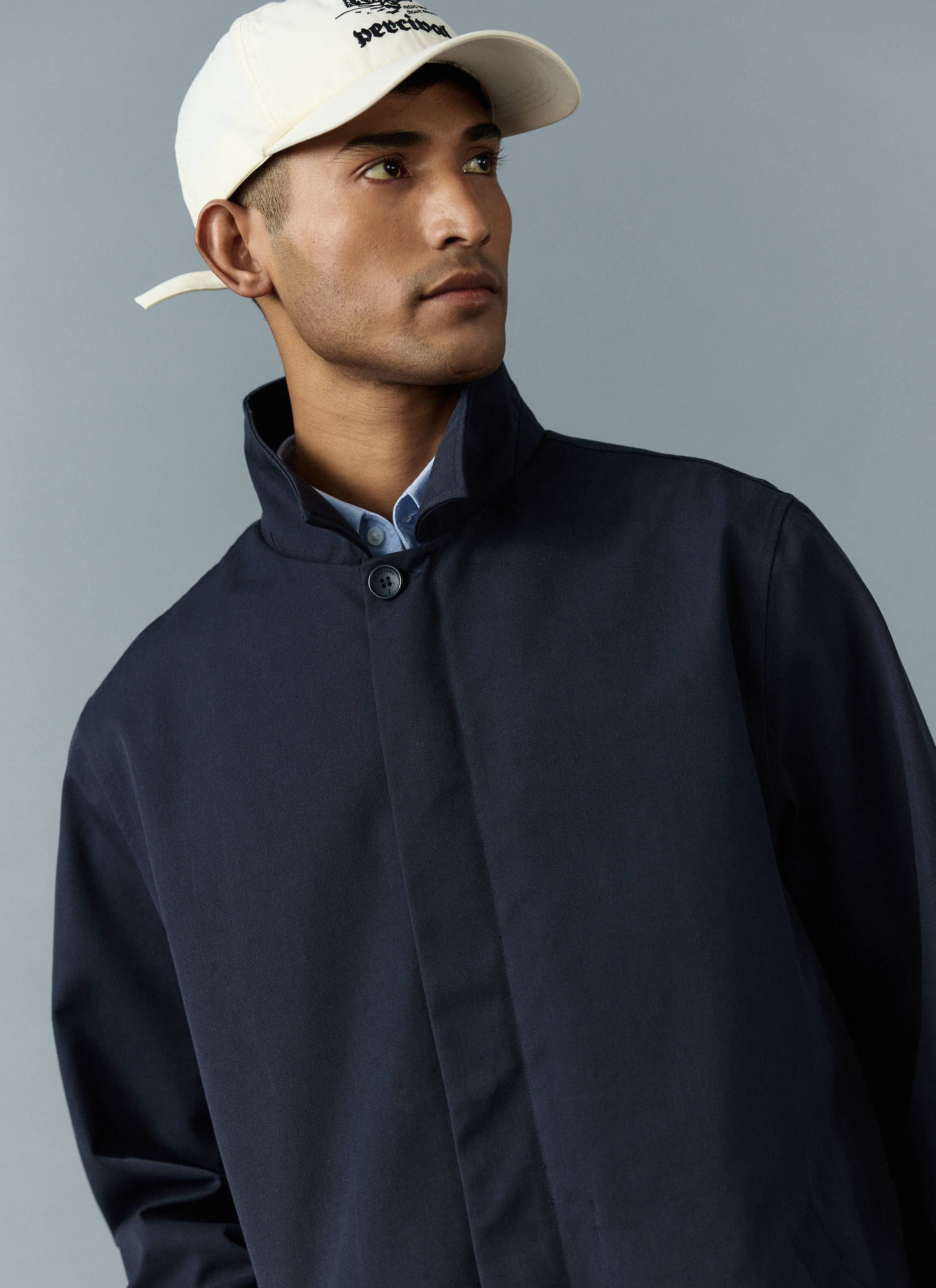 A close-up of a model wearing the navy Greyson Long Trench Coat from Percival with a cream branded baseball cap.