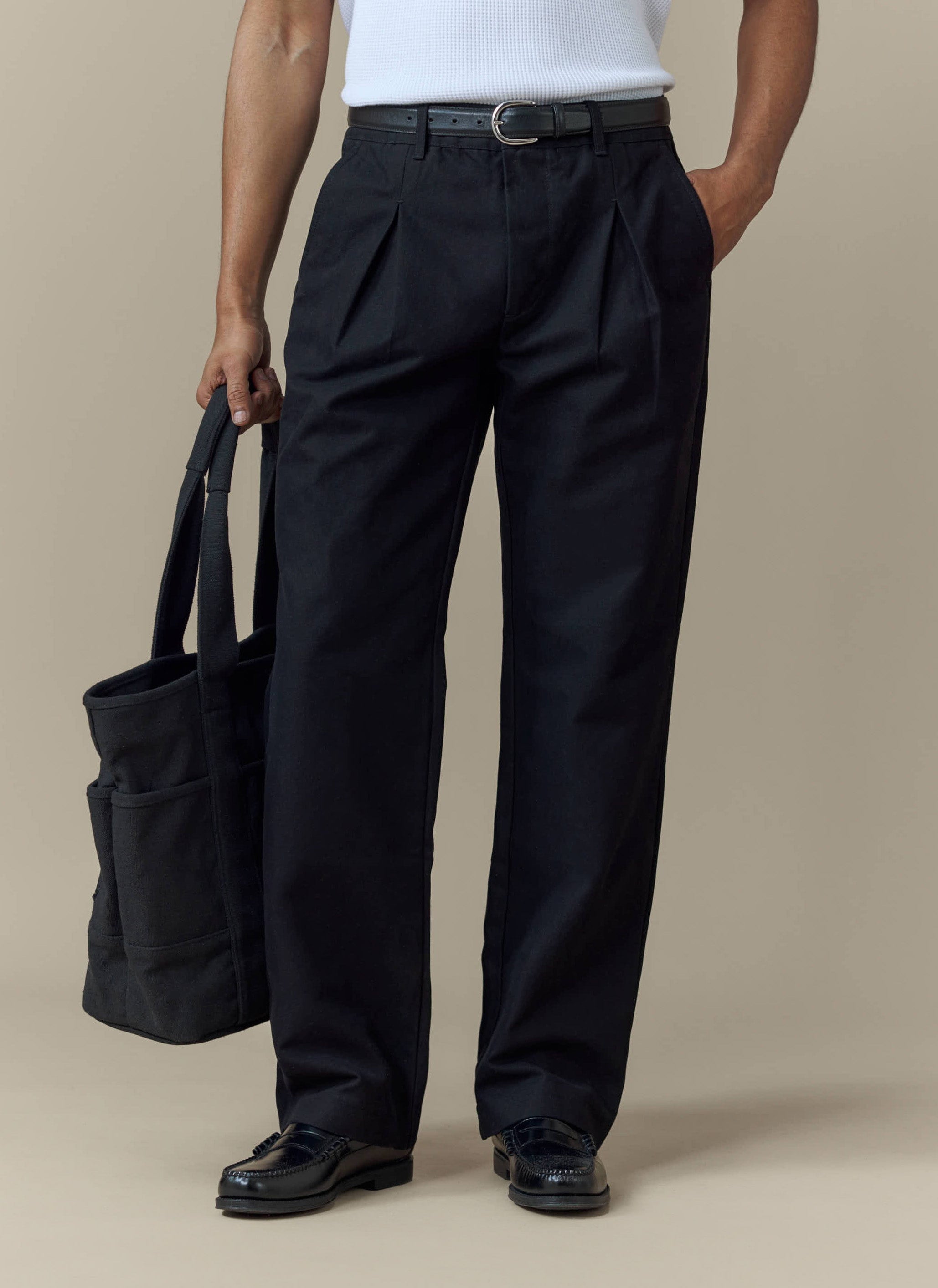 A model wears the Percival Seb Inverted Pleat Trousers in black, styled with a white vest, loafers and a tote bag.
