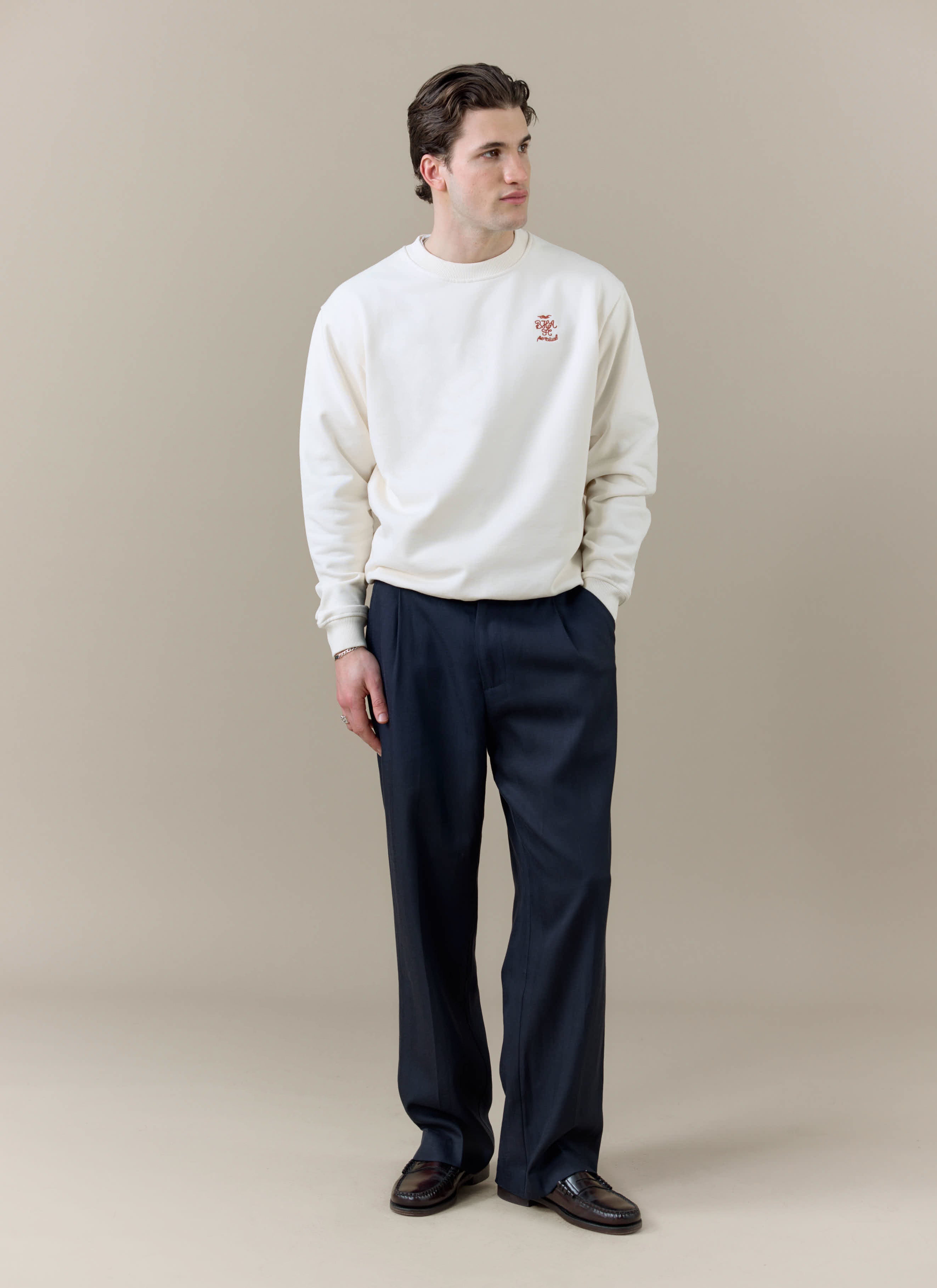 A model wears the cream Percival x Brighton & Hove Albion FC sweatshirt with dark smart trousers against a neutral backdrop.
