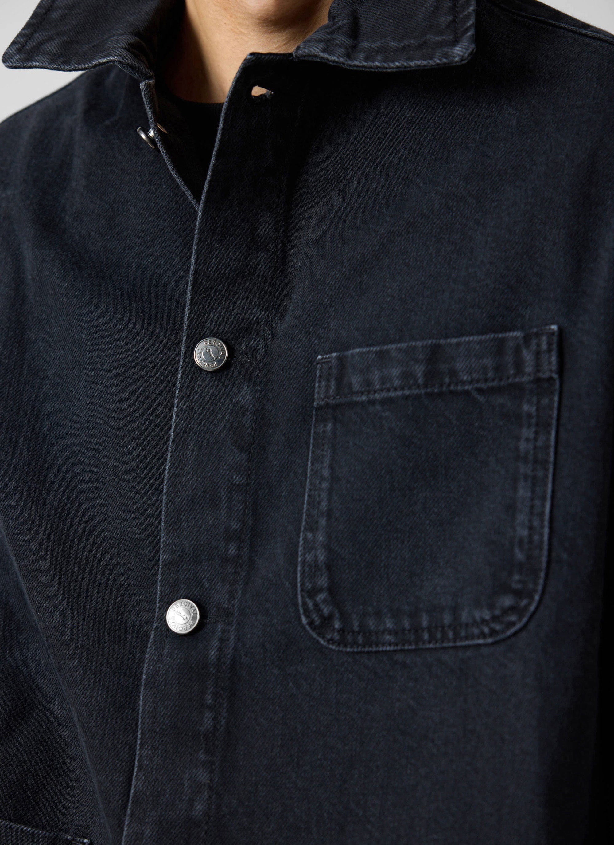 A close-up of the Percival Gove Chore Jacket in black denim, showing the collar, chest pocket, and branded buttons.