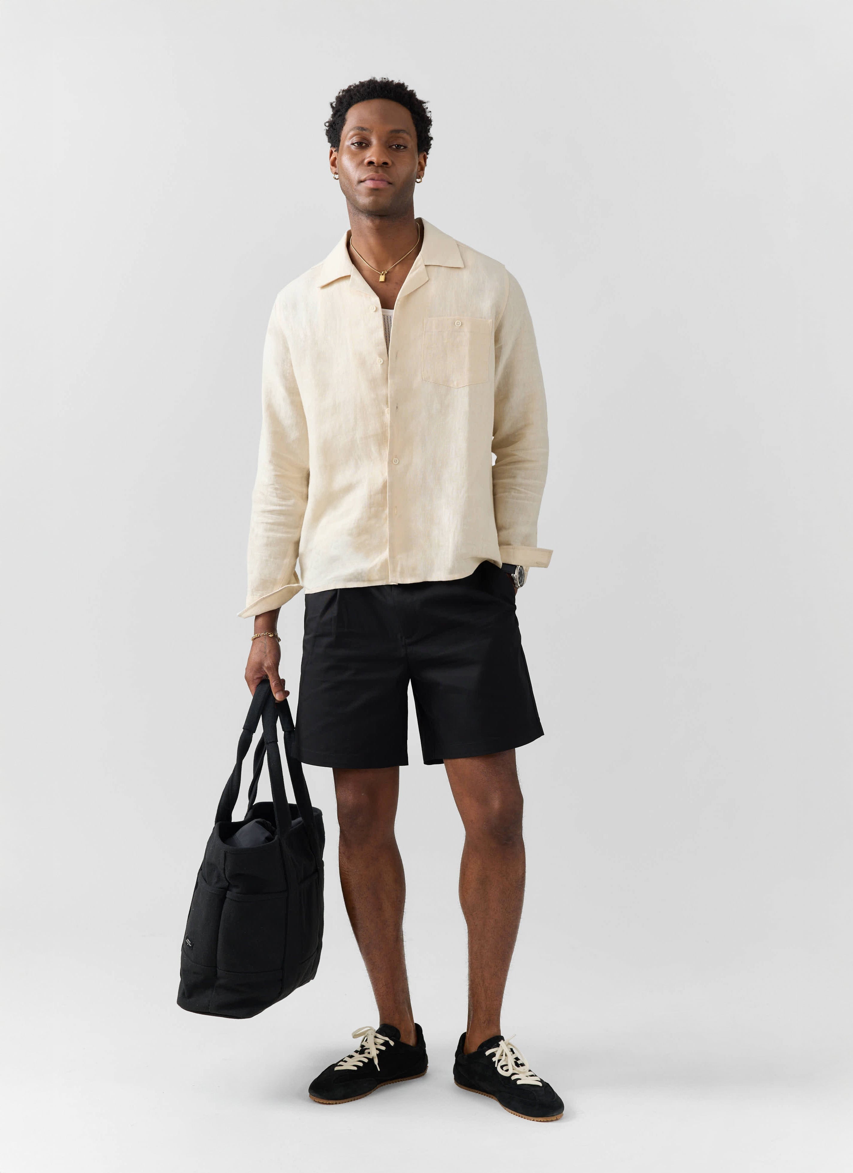 A model wears the Percival Long Sleeve Cuban Linen Shirt in cream with black shorts and a tote bag.