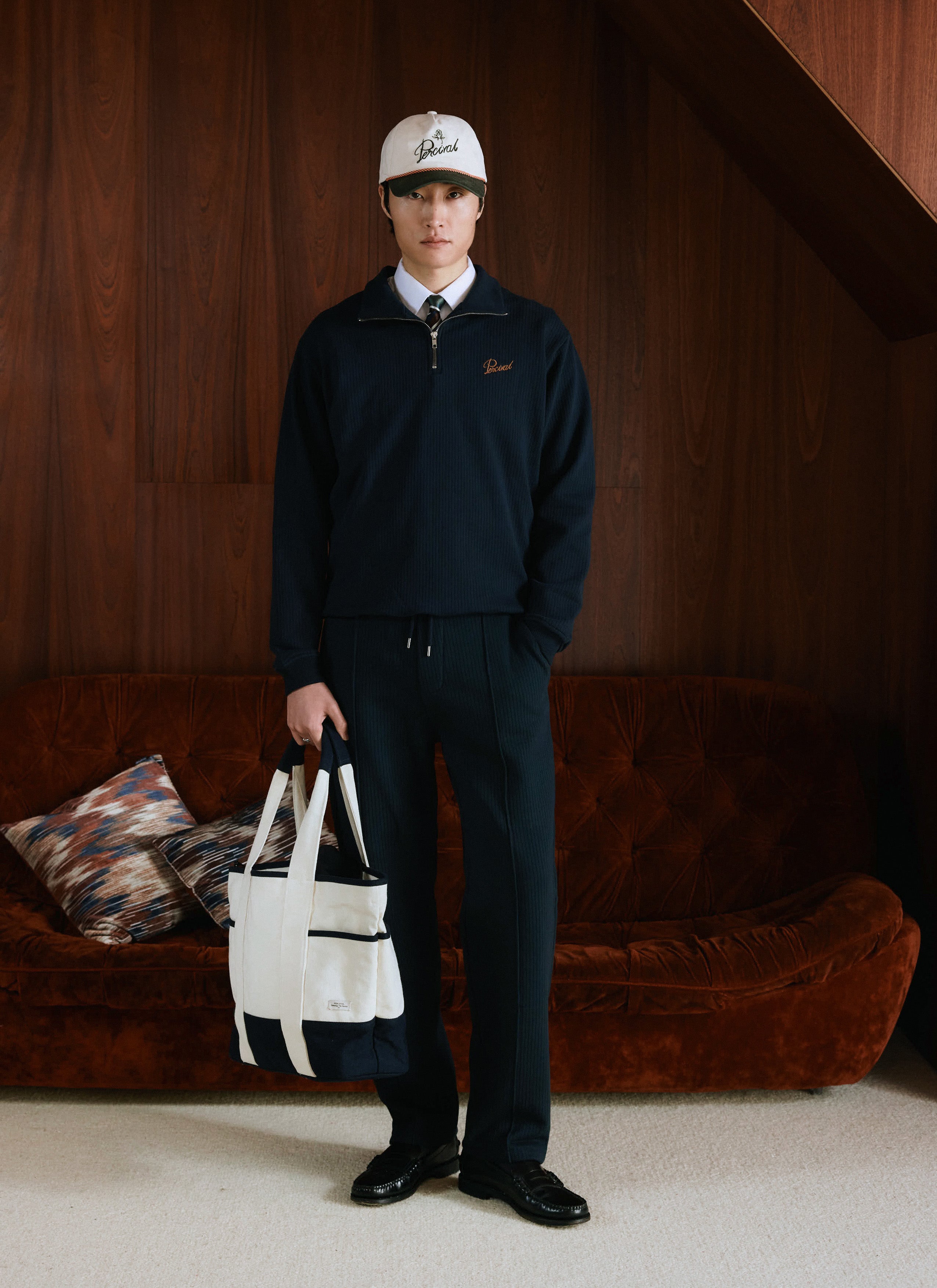 A model wearing the navy Percival cotton funnel neck 1/4 zip jumper with matching trackpants, a cap, and holding a tote bag.