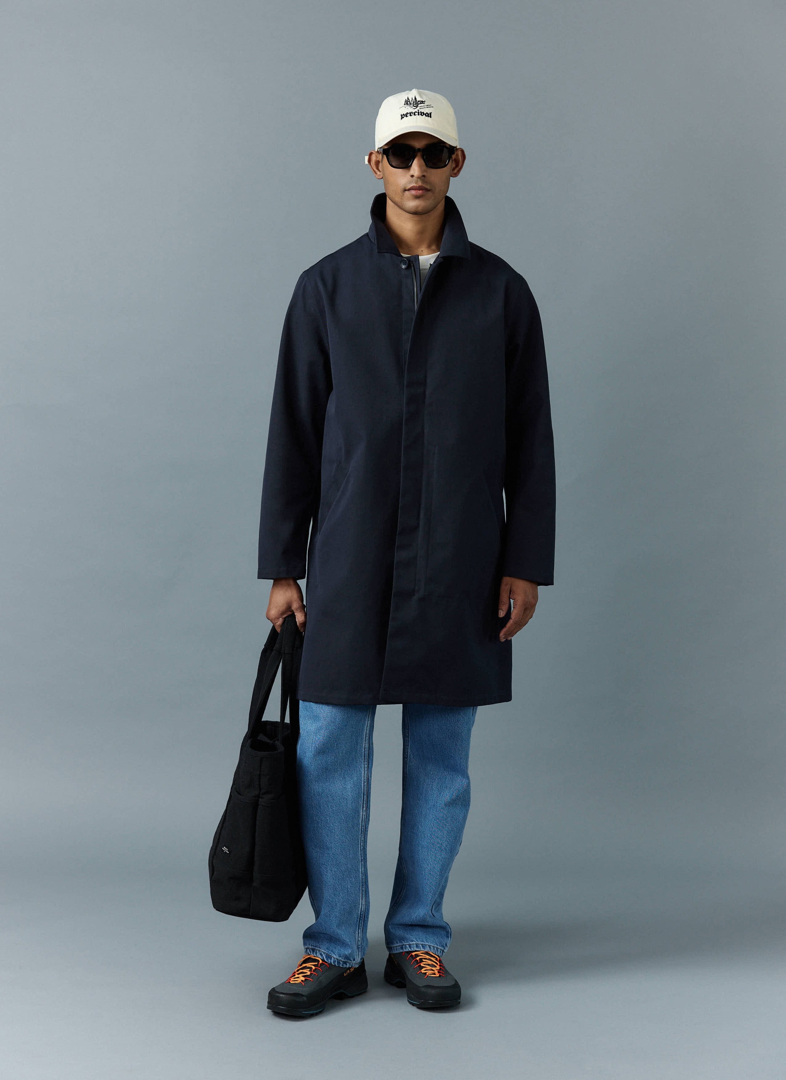 A model in a studio wears the Percival Greyson Long Trench Coat in navy with blue jeans, a white cap, and a black tote bag.