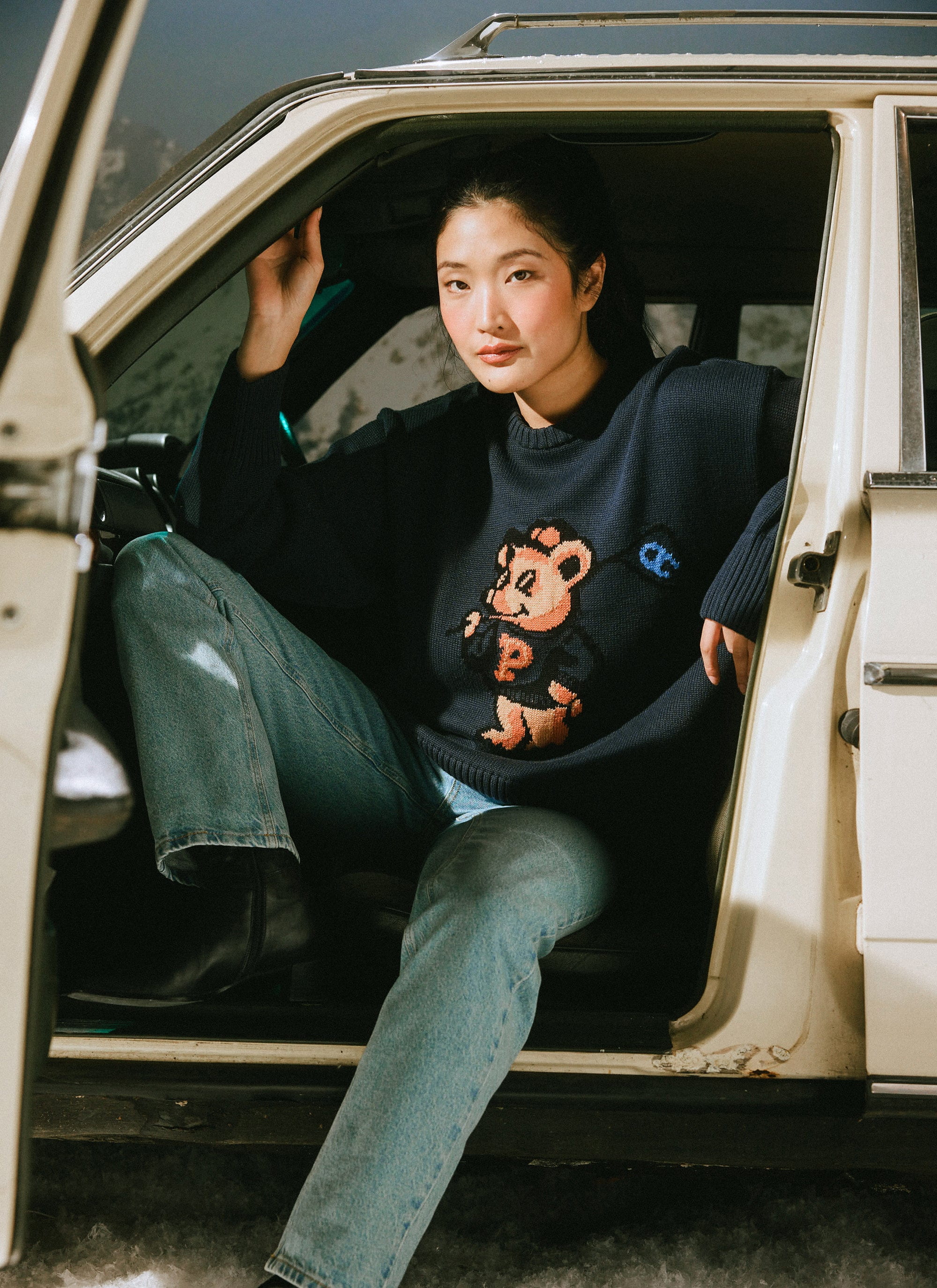 A model wears the navy Champion x Percival Jacquard Bear crew neck jumper, sitting in the open door of a vintage car.