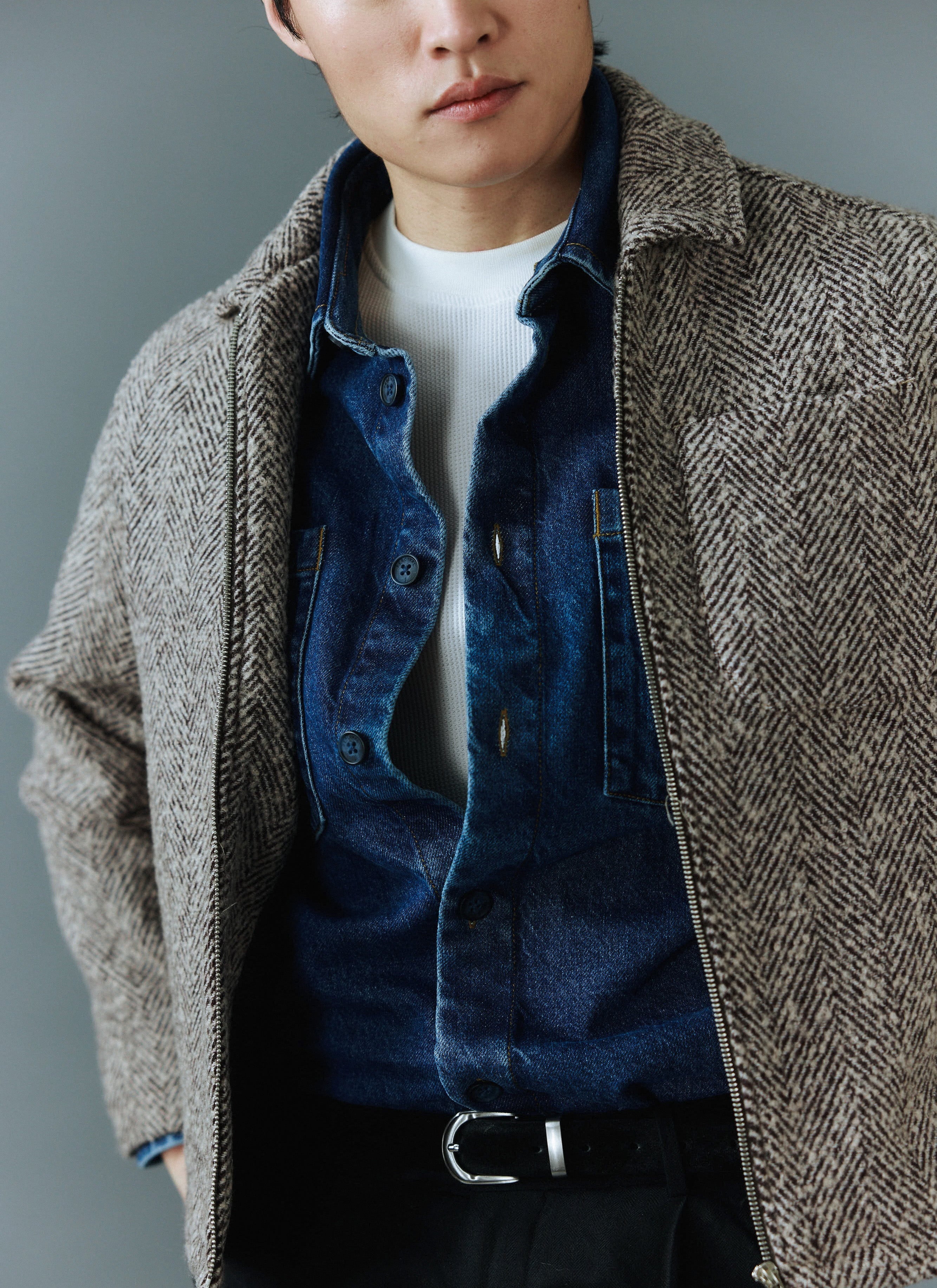 A close-up of a model wearing the Percival Herringbone Zip Jacket in ecru, styled open over a denim shirt.