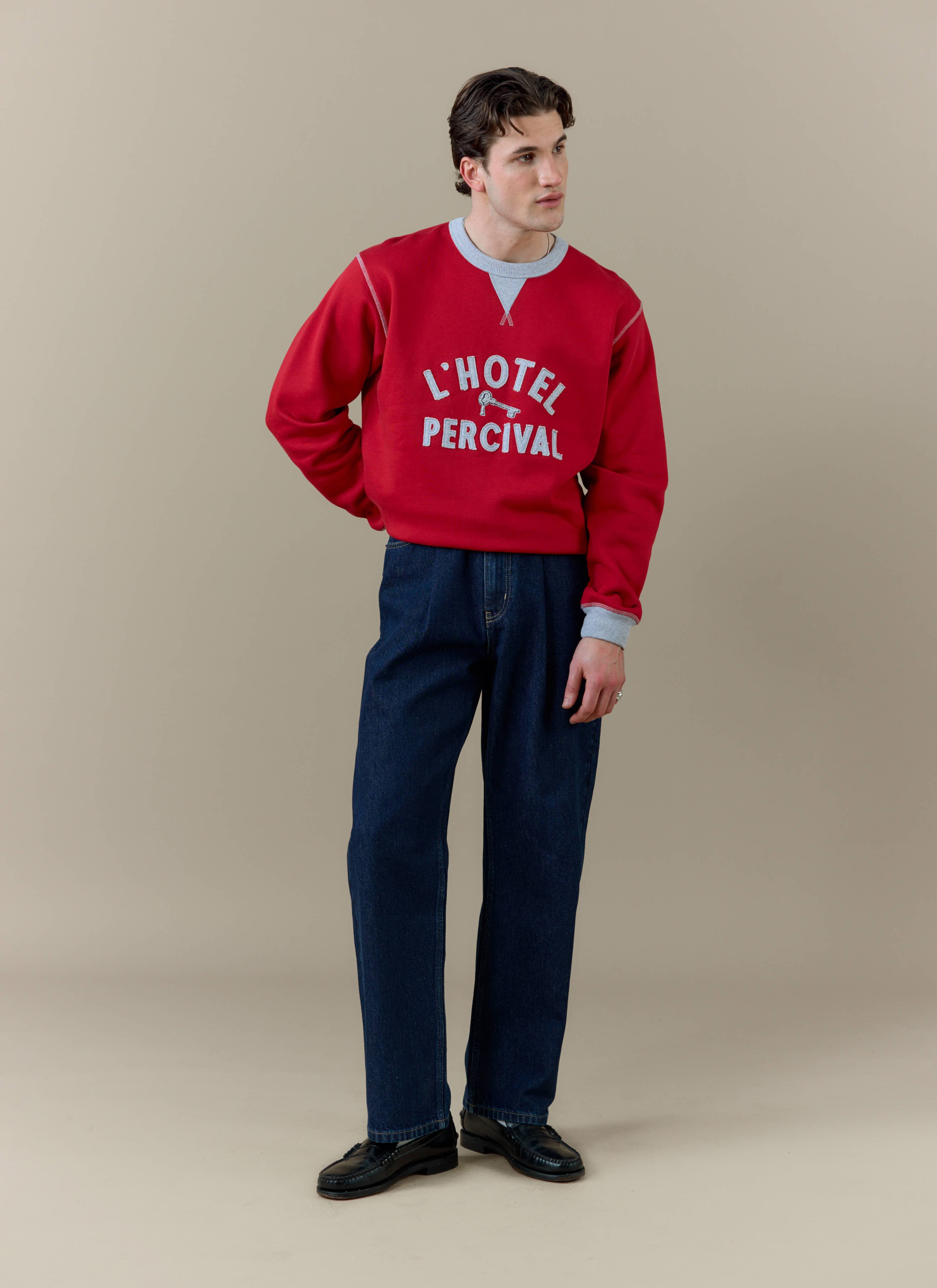 A model wearing the red Ronald sweatshirt from Percival with 'L'Hotel Percival' applique and dark blue trousers.