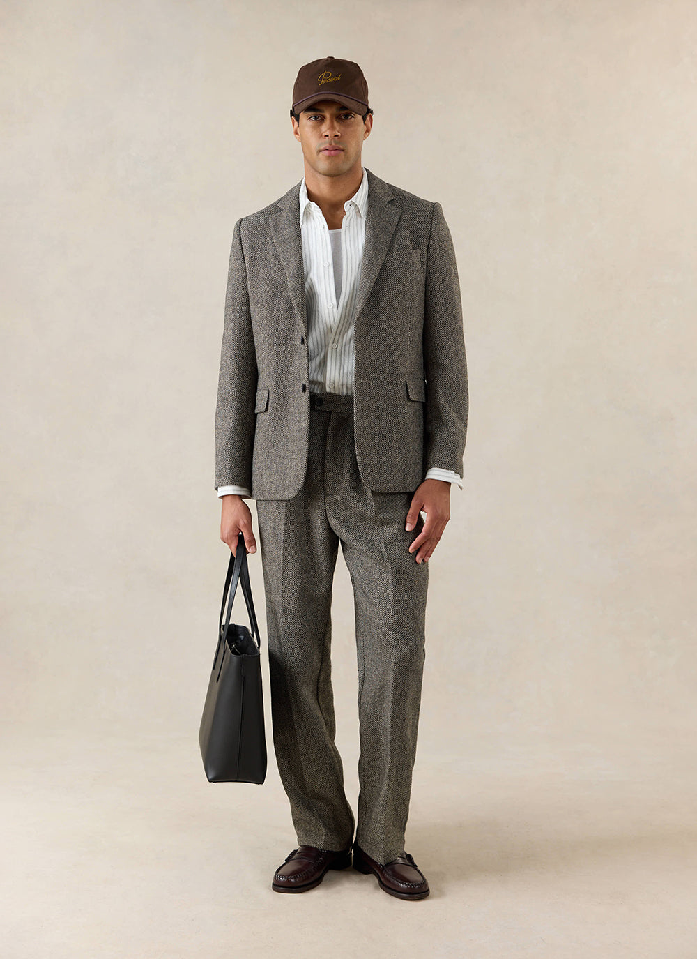 A model wears the Percival Tailored Blazer in Birdseye Wool Grey, styled with matching trousers, a brown cap and black bag.