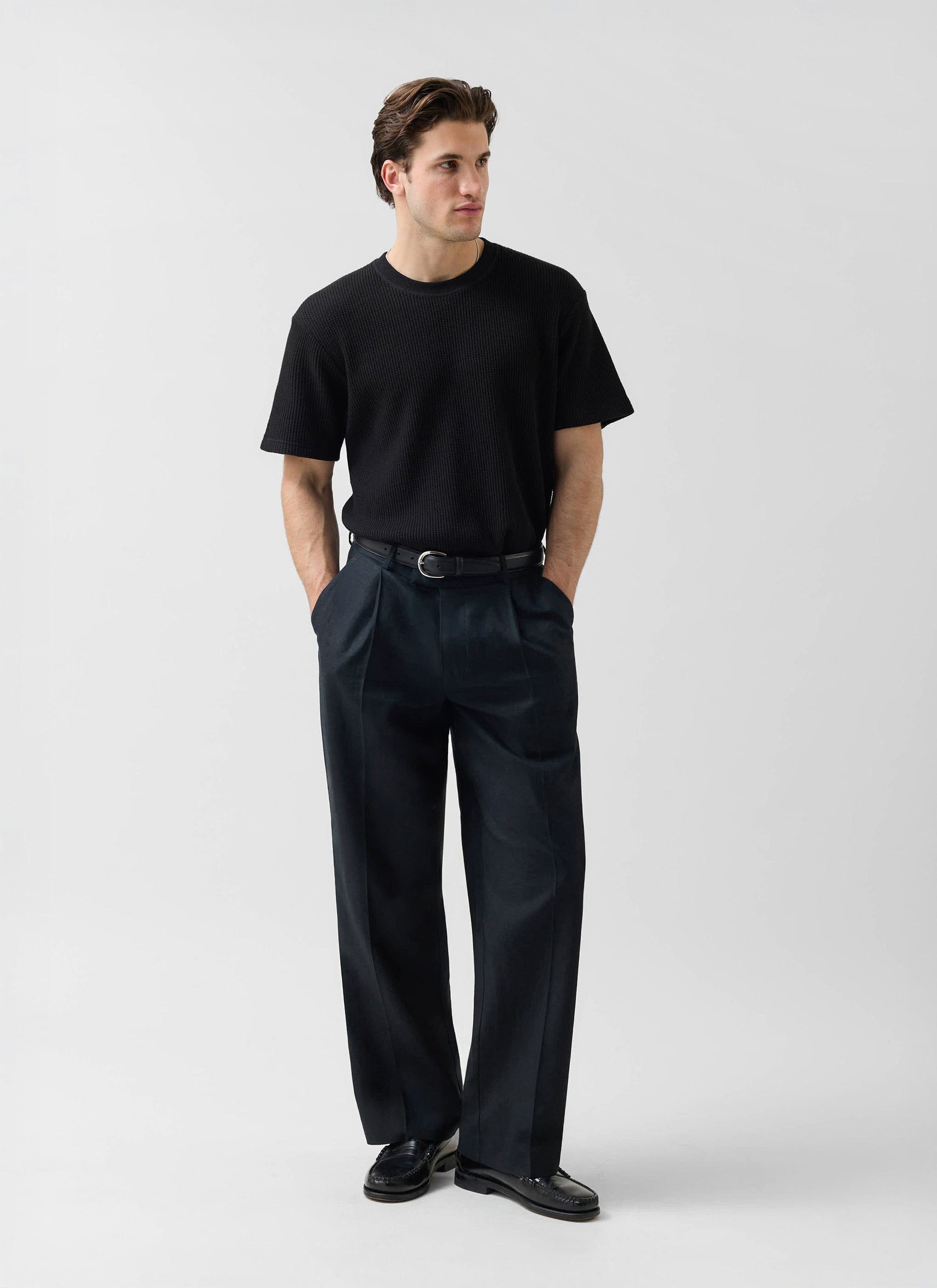 A model wears the black Wide Leg Linen Trousers from Percival with a black t-shirt and loafers against a white background.