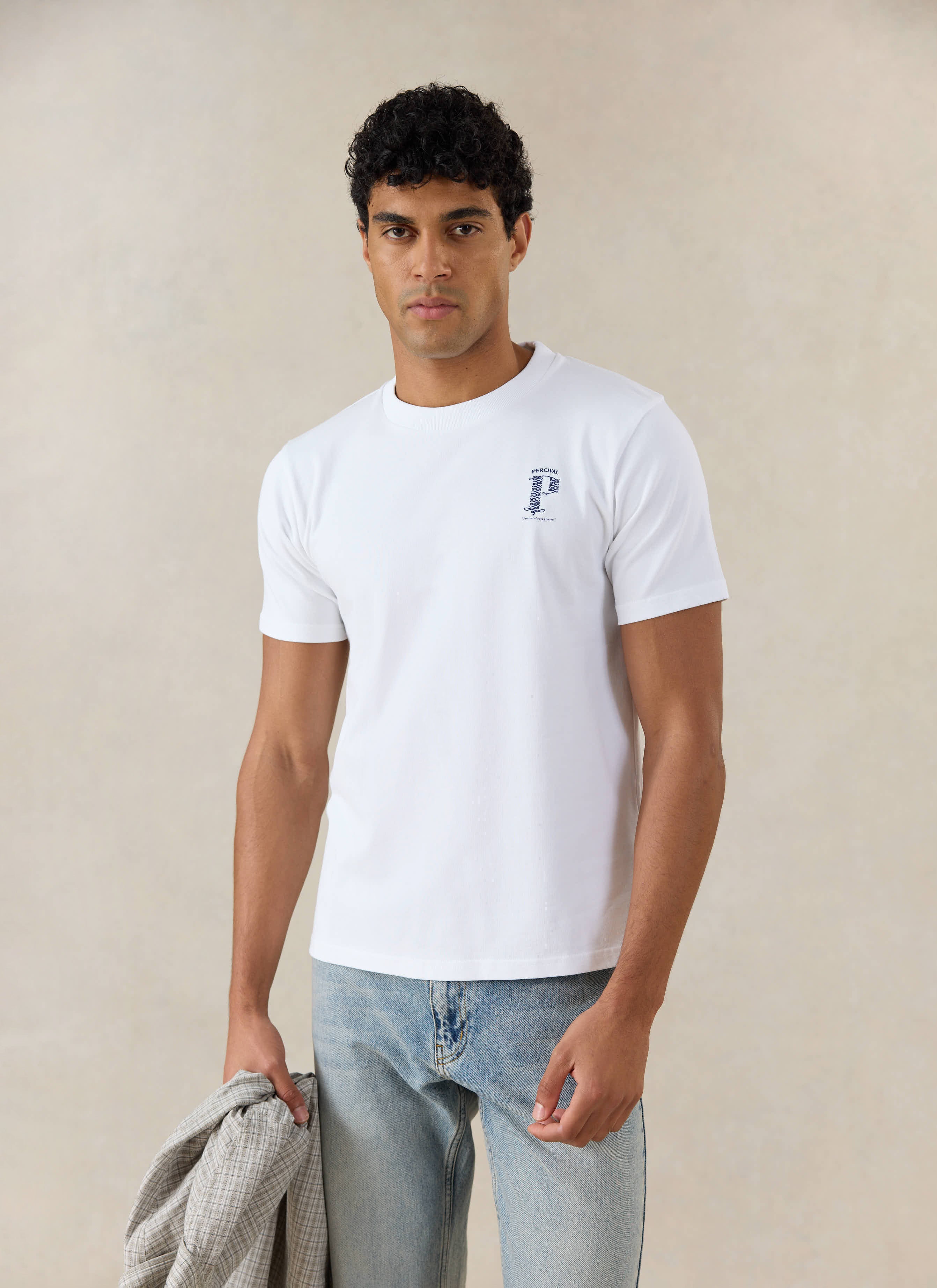 A model wears the white Percival Cyril T-shirt with a blue graphic, in light blue jeans and holding an overshirt.