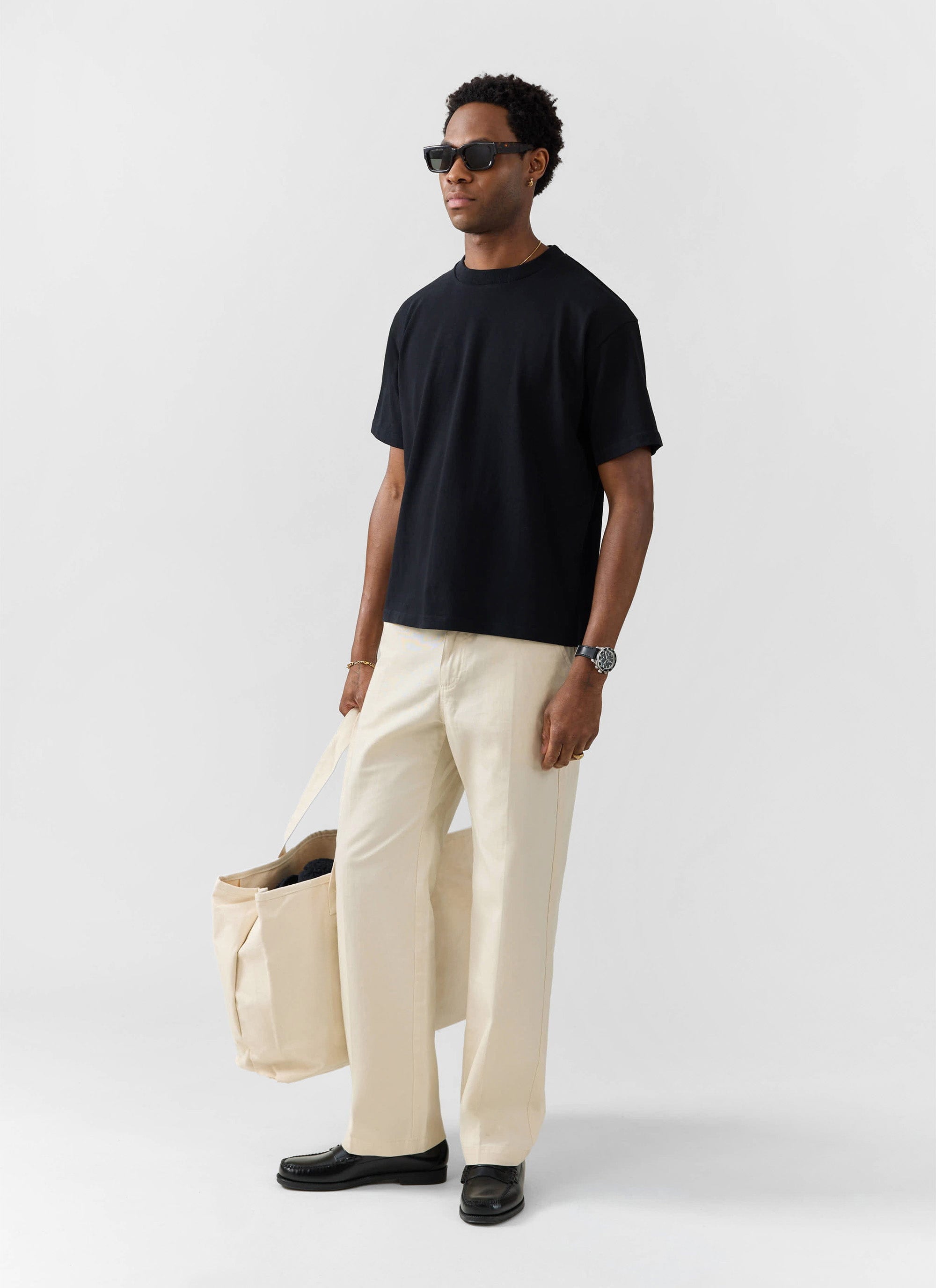 A model wears the Percival oversized t-shirt in black organic cotton, styled with cream trousers and black sunglasses.
