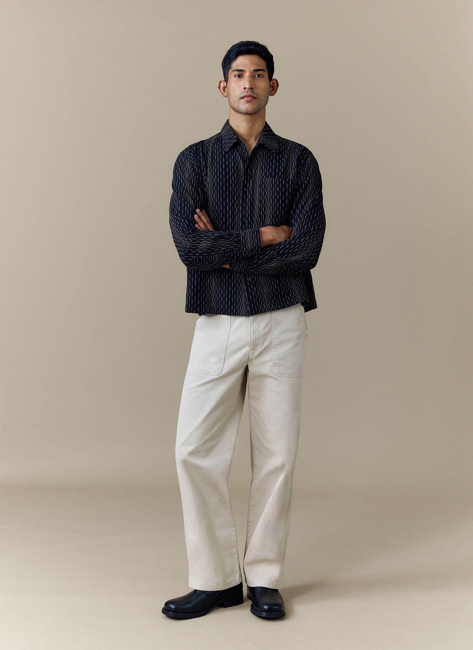 A model stands with arms crossed wearing the black Leland Cuban Boxy Shirt by Percival, styled with off-white trousers.