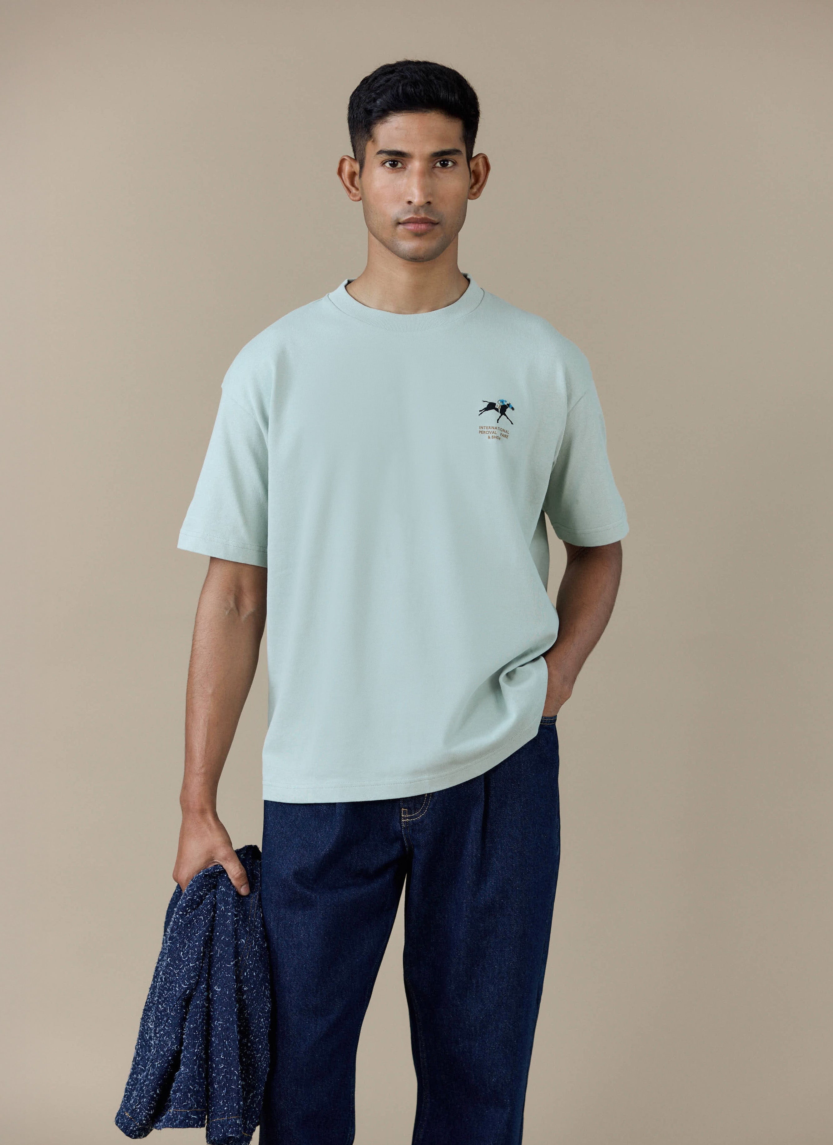 A model wearing the Percival Fair and Show Oversized T Shirt in sage green with the chest graphic.