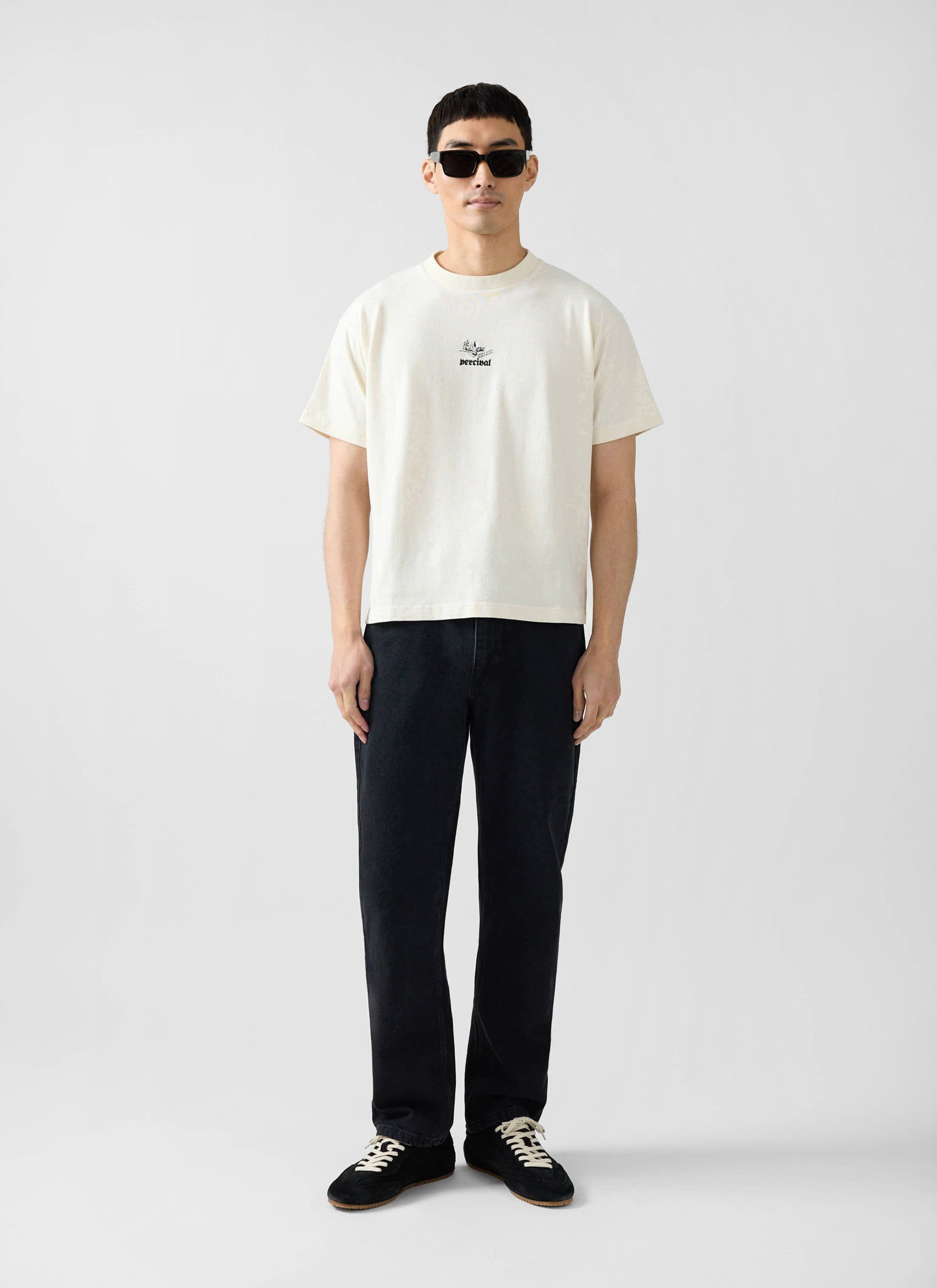 A model wears the Percival Wildlands Oversized T Shirt in ecru with black trousers and sunglasses on a white background.