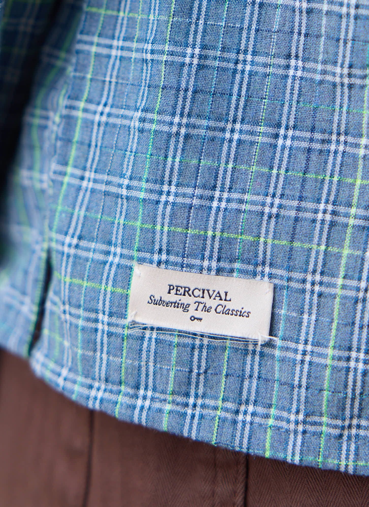 Close-up of the Percival Hinsley Checked Boxy Shirt in blue cotton, showing the white woven brand label at the hem.