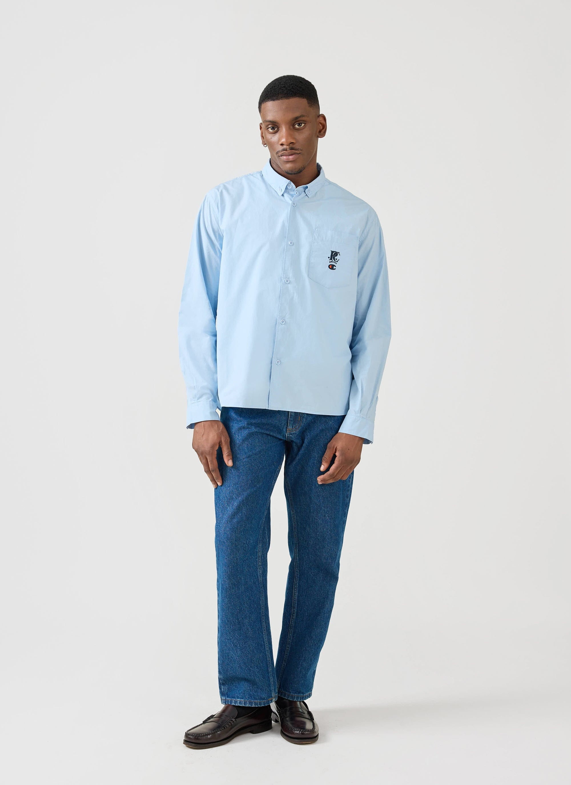 A model wears the light blue Champion x Percival Relaxed Oxford Shirt with blue jeans in a full-length Percival studio shot.