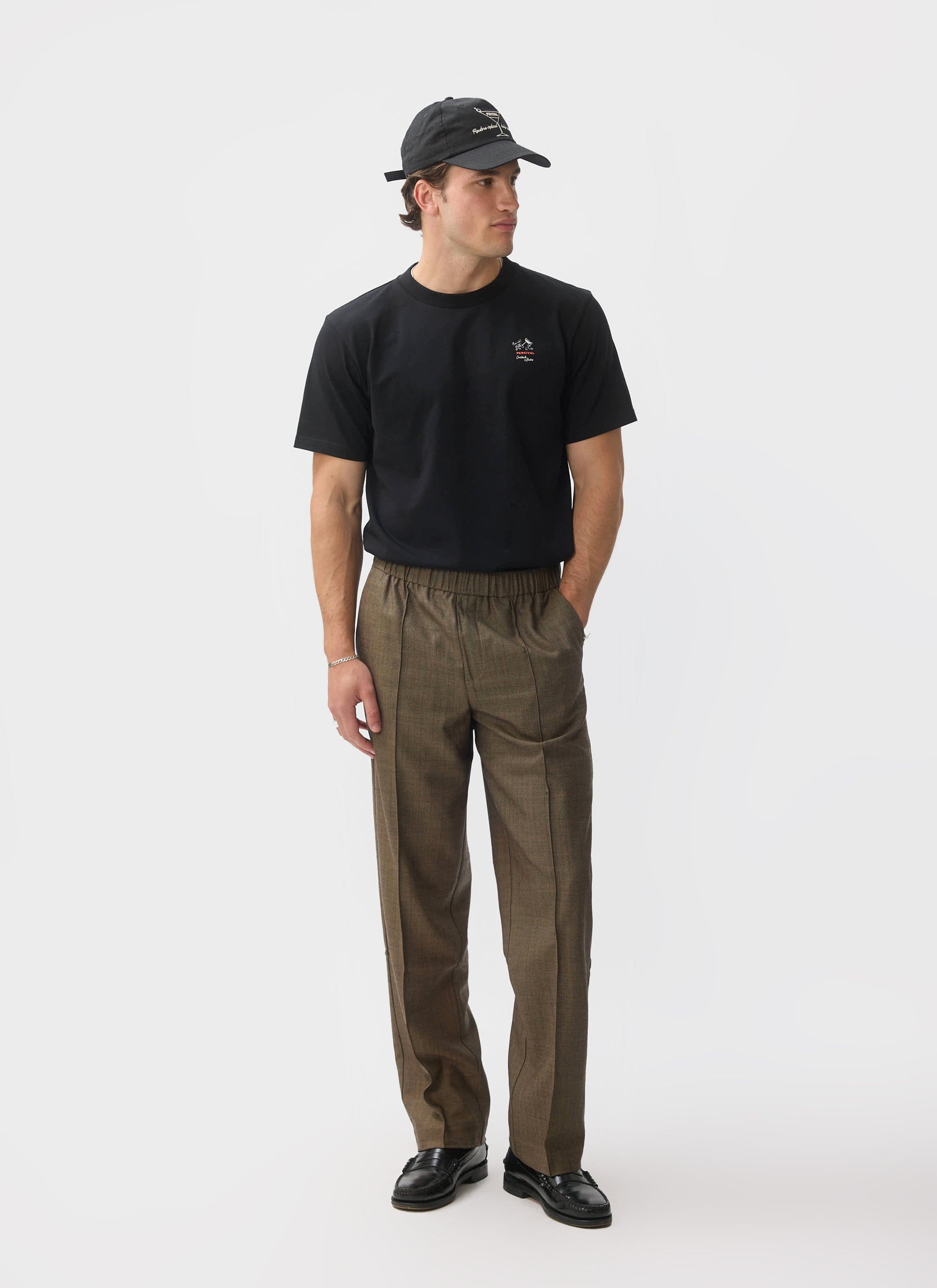 A model wears the black organic cotton À La Carte T Shirt from Percival, styled with brown checked trousers and a black cap.