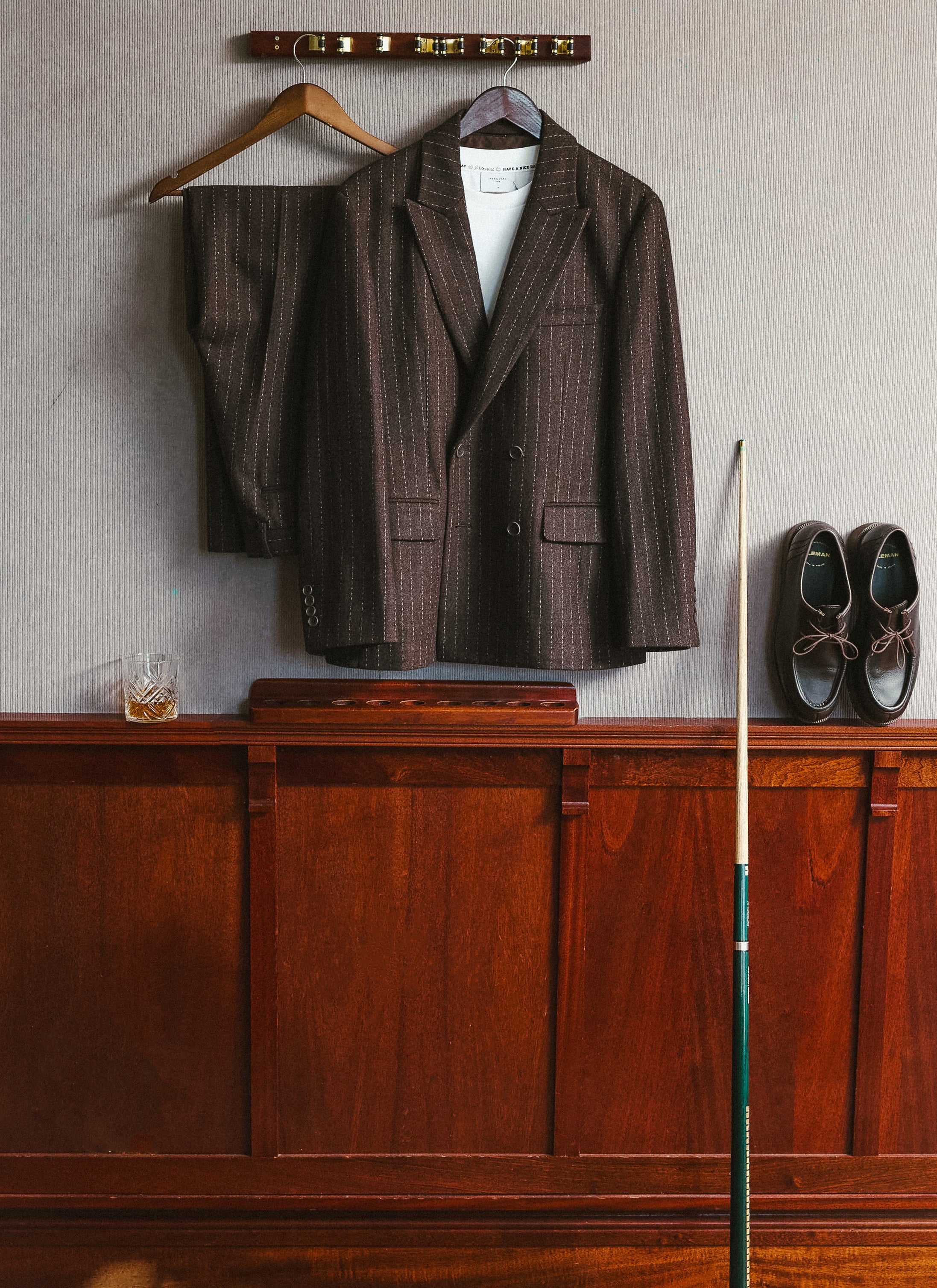 The Percival espresso wool pinstripe suit hanging on a wooden rack alongside matching brown leather shoes.