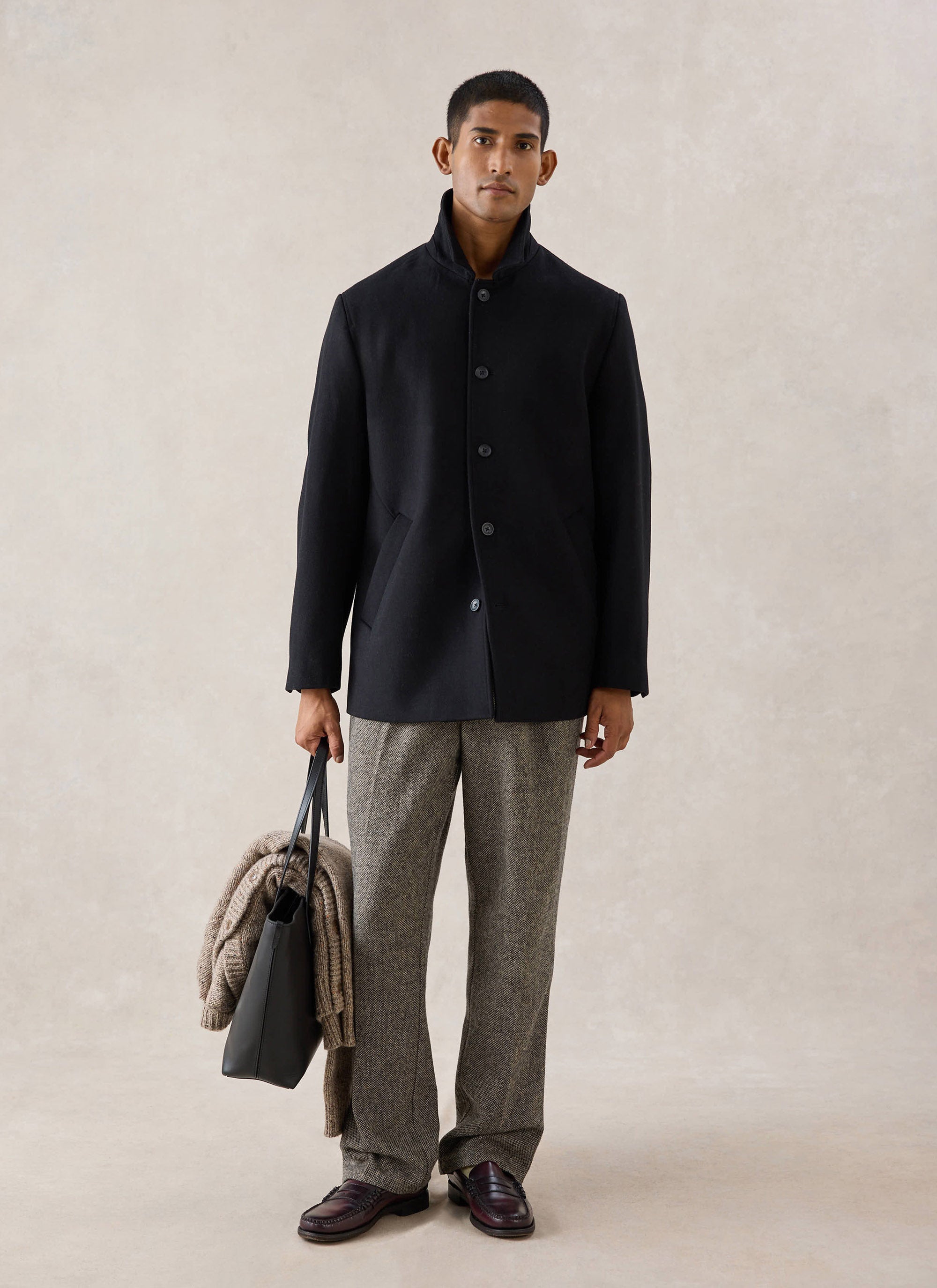 A model wears the black melton wool peacoat from Percival over patterned trousers, holding a black leather bag in a studio.