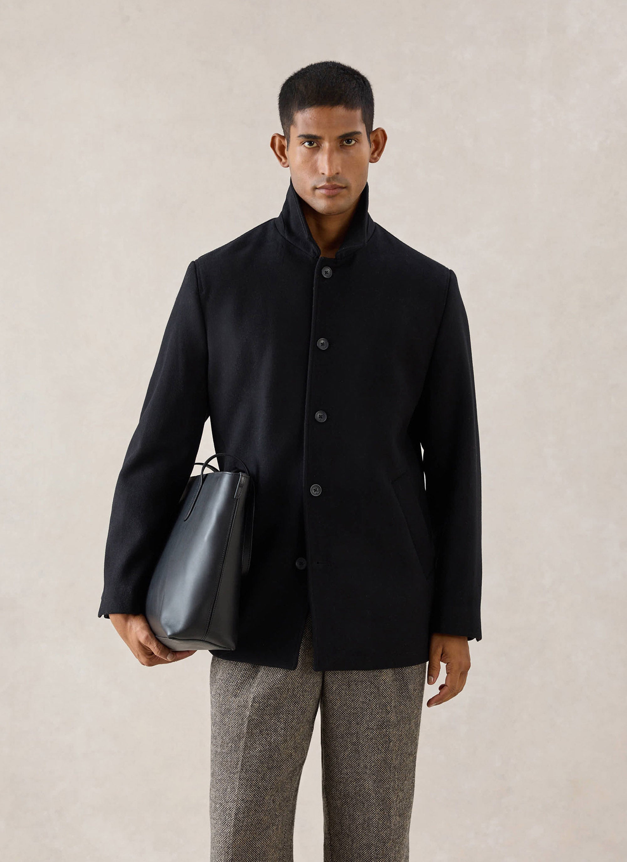 A model wears the black Melton wool peacoat from Percival with a popped collar, holding a black leather bag in a studio.
