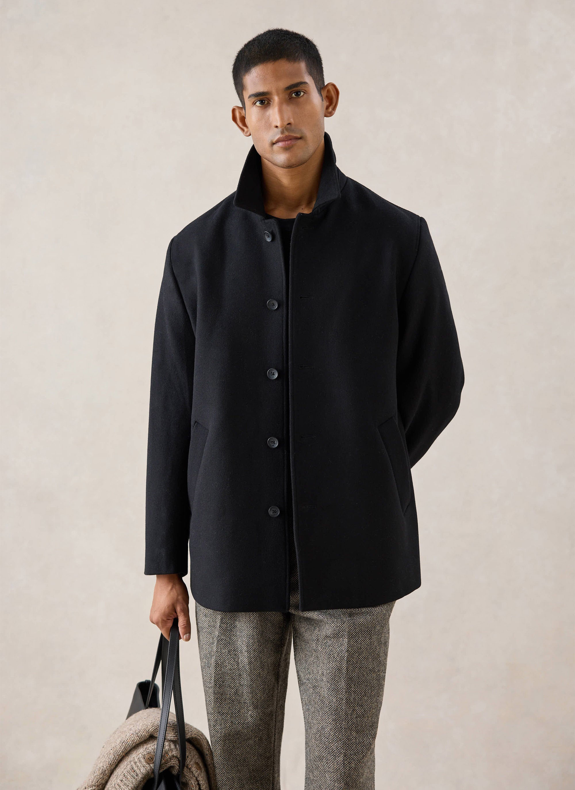 A model wears the black melton wool peacoat from Percival over patterned trousers while holding a bag in a studio setting.