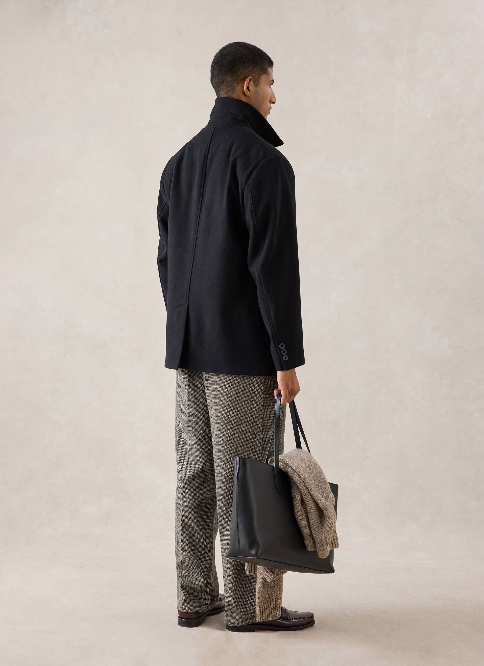 Rear view of a model wearing the black Melton wool peacoat from Percival, holding a black tote bag.